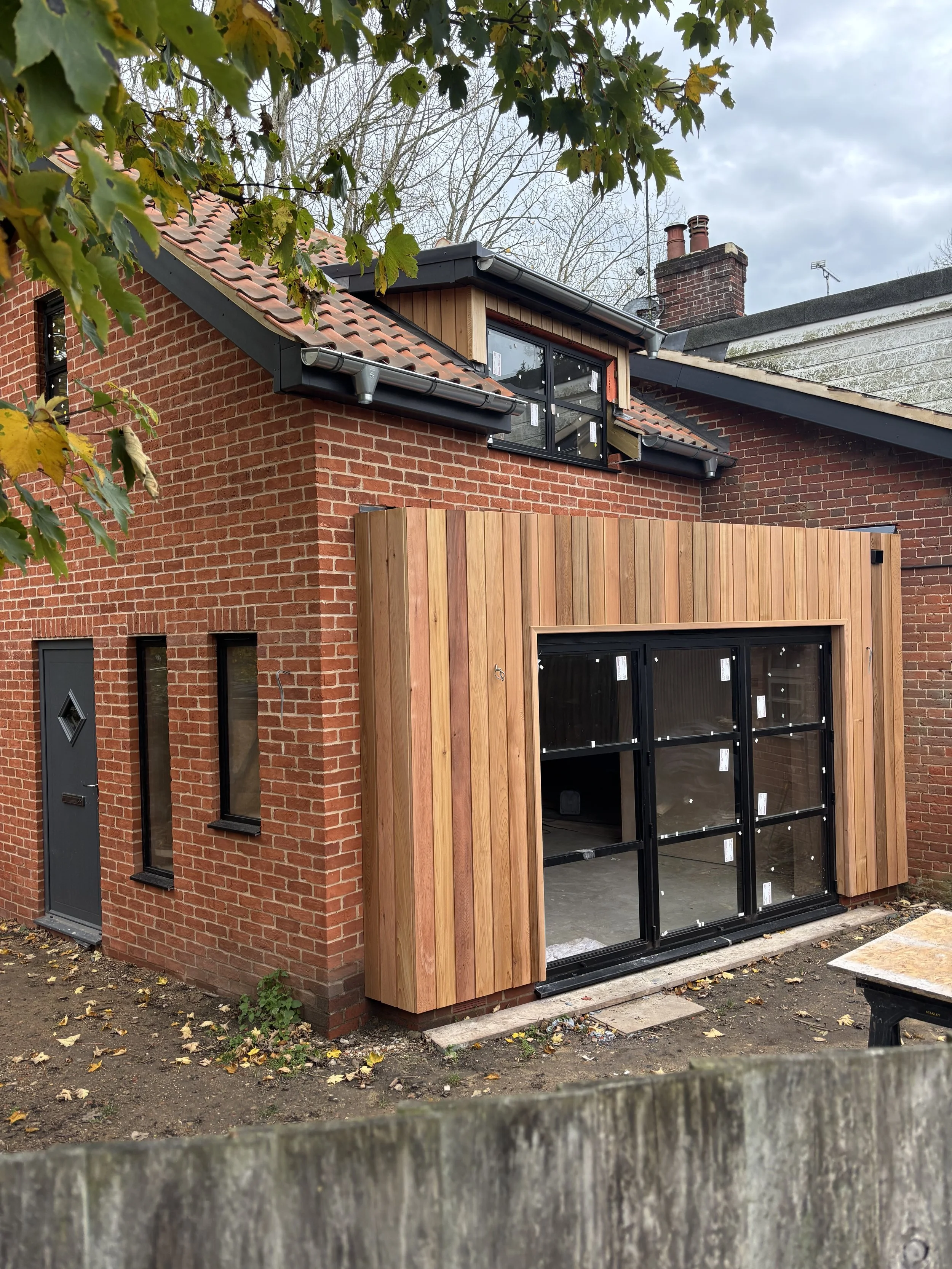 A brick house with a wooden addition under construction. The addition has large black-framed glass doors and vertical wooden siding. The house has a red-tiled roof, with some trees in the background and a cloudy sky.