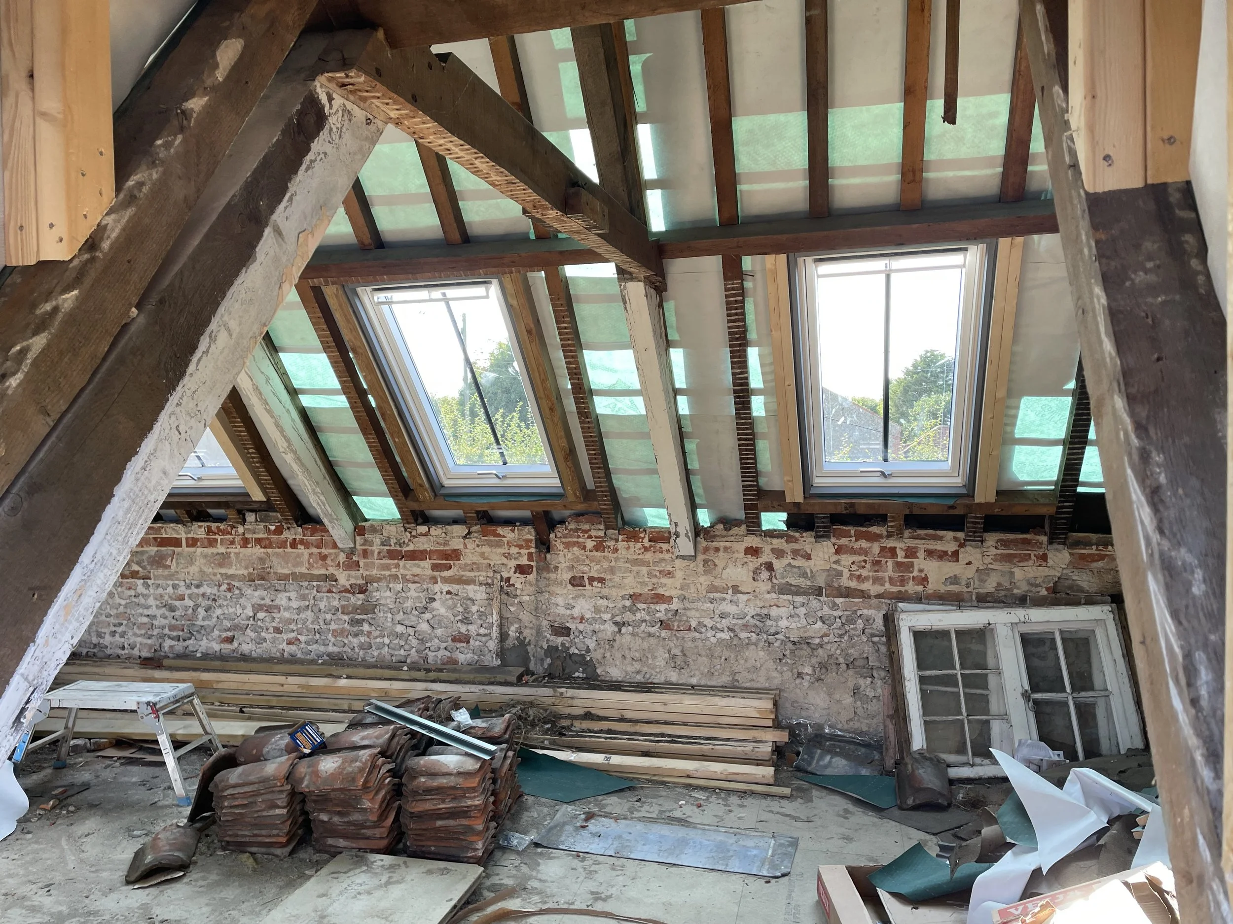 Interior of a building under construction with exposed wooden beams, brick wall, large windows, and construction materials scattered on the floor.