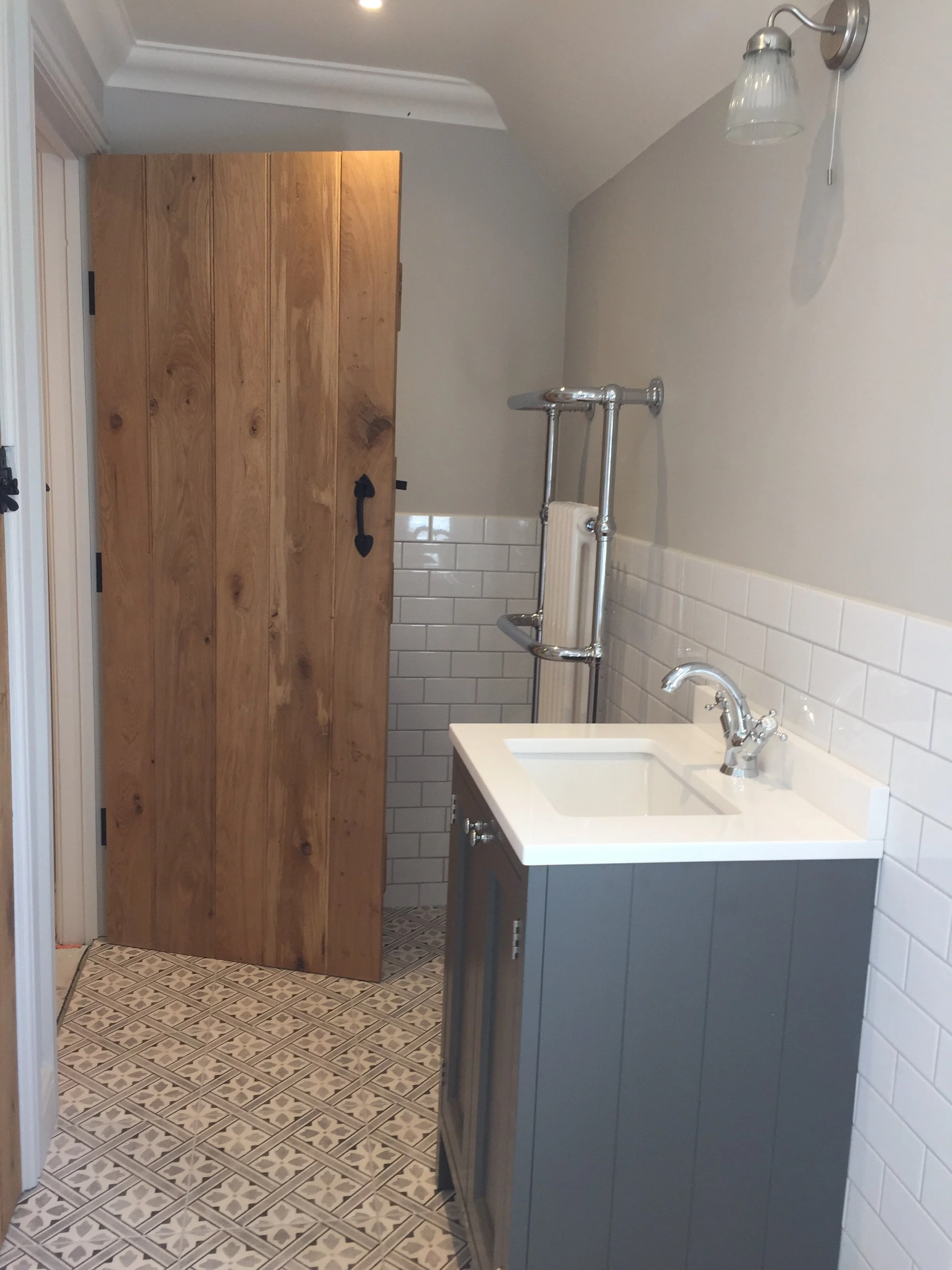 Small bathroom with patterned floor tiles, a wooden door obscuring part of the space, a modern sink with a gray cabinet, a small radiator, white tiled wall, and a wall-mounted light fixture.