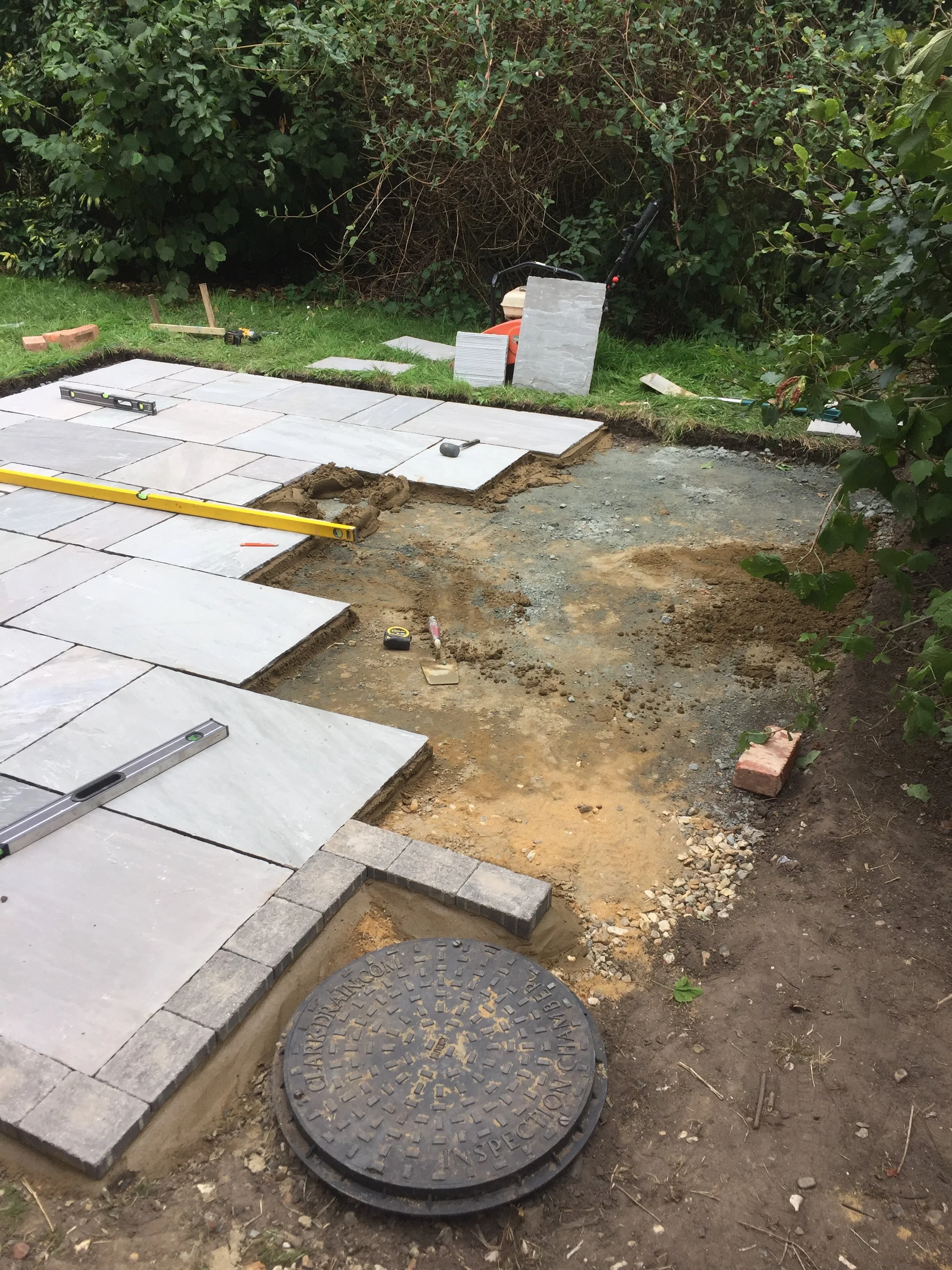 Construction site where paving stones are being installed on a pathway. There are tools and building materials scattered around, with some stones already laid and others awaiting placement. The area is partially excavated, with dirt and gravel visibl