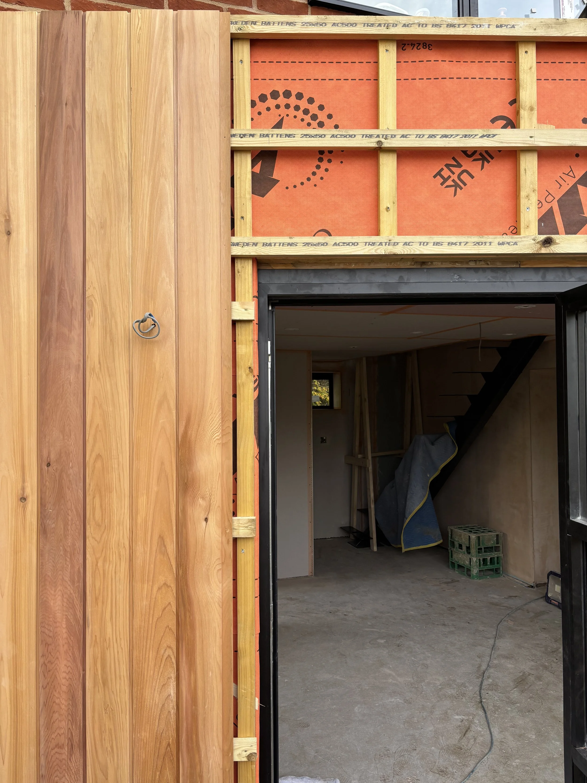 Construction site showing a partially built wall with wooden framing and exterior siding, with an interior space visible through a doorway.