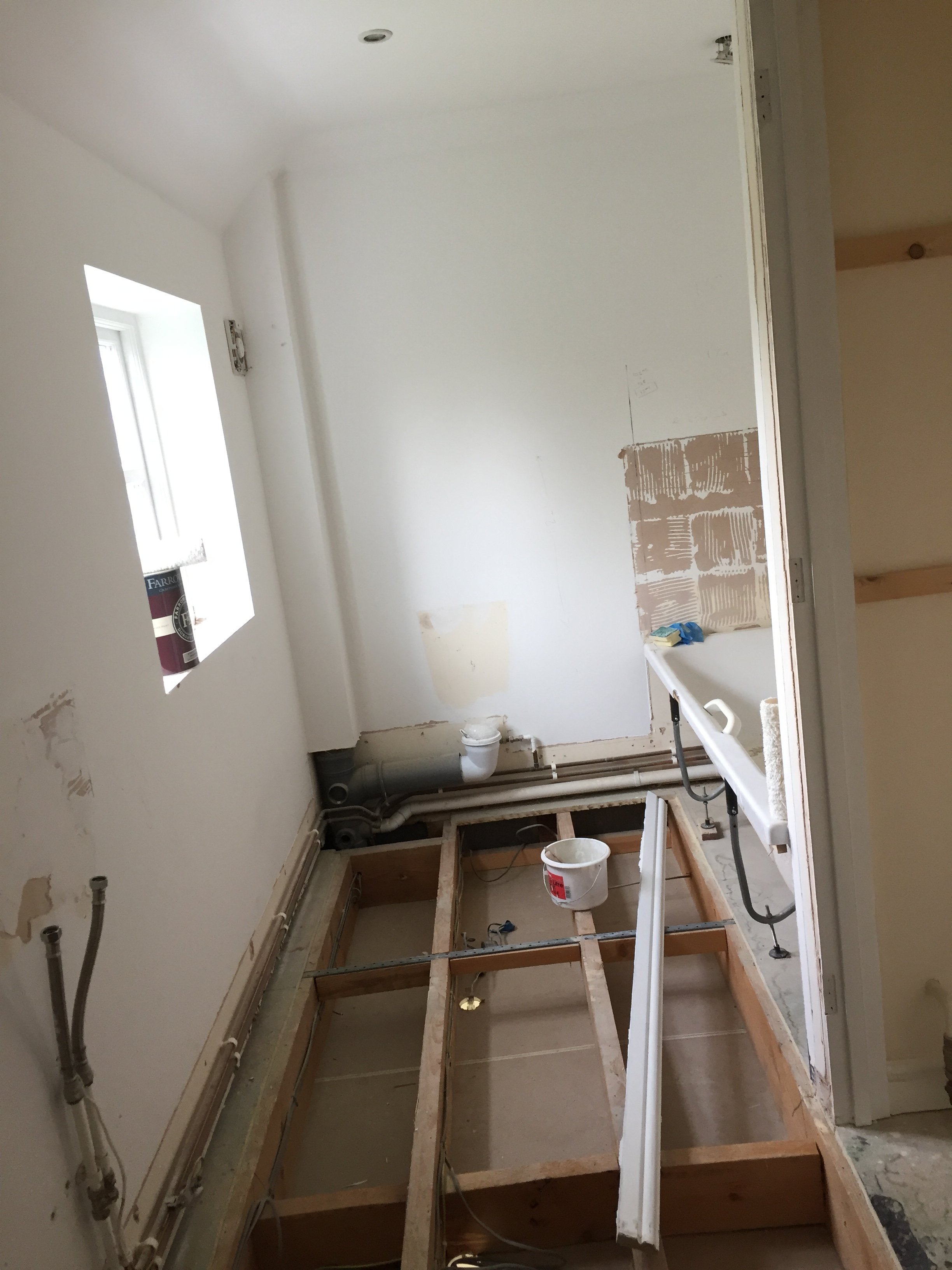 A room under renovation with plumbing pipes, exposed wooden framework, and a partially removal of flooring. The walls are white, and there is a small window on the left side of the room.