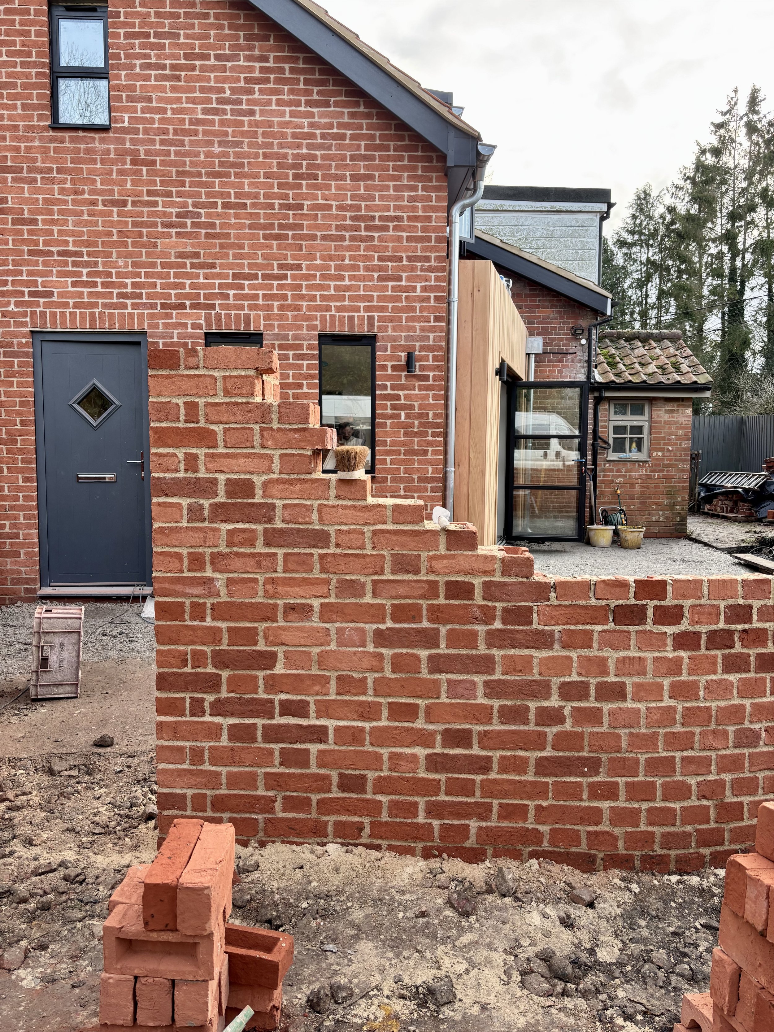 A construction site showing a red brick wall with a small brick ledge, neighboring brick building with black window frames, a black door, and unfinished ground area with construction materials and scattered bricks.