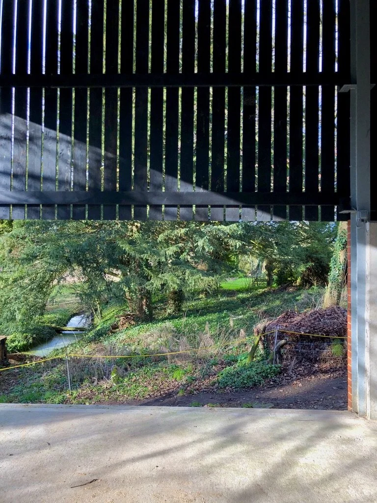View through an open structure with a slatted roof, looking out onto a lush green landscape with trees, grass, and a small stream.