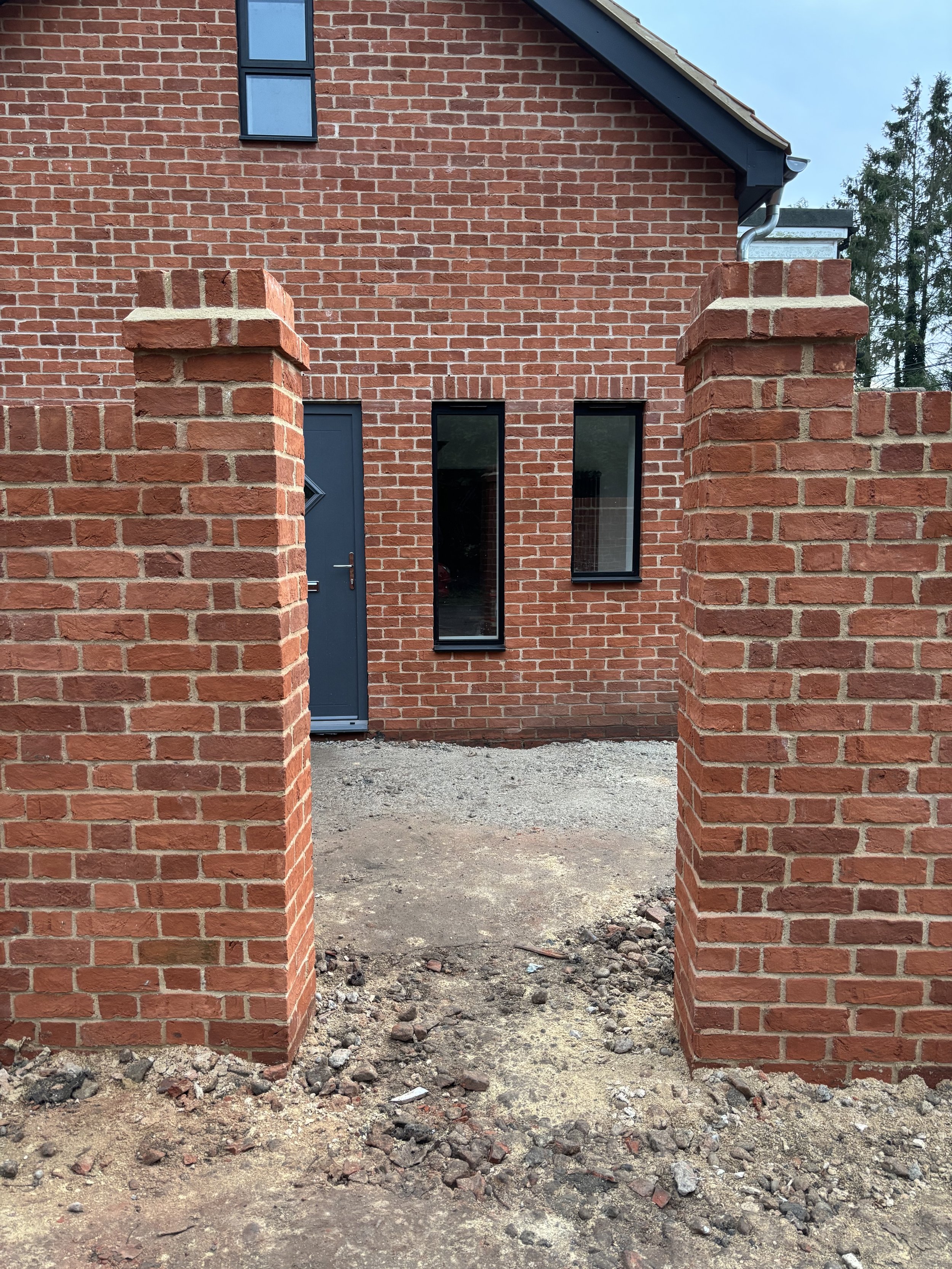 Building under construction with a brick wall, open space, and two brick pillars in the foreground. The ground is rough with dirt and bricks scattered.
