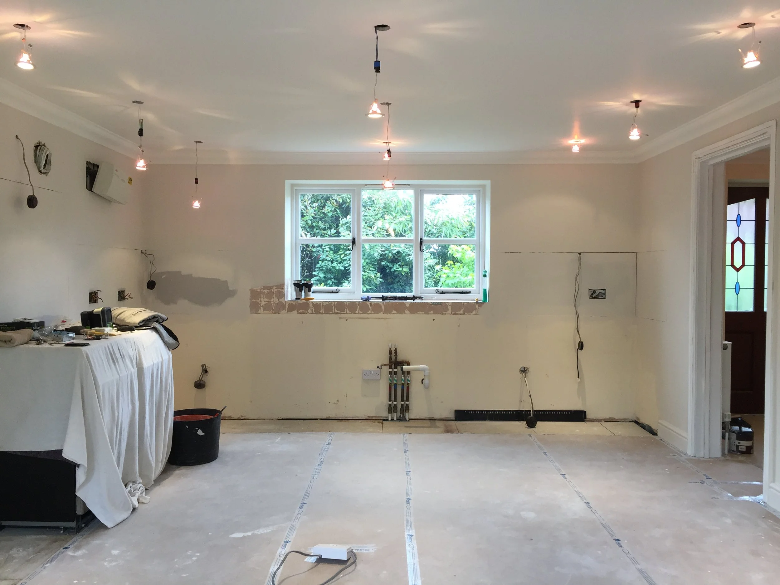 A living room under renovation with unfinished walls, a large window with green foliage outside, and hanging light fixtures from the ceiling. Tools and construction materials are scattered around.