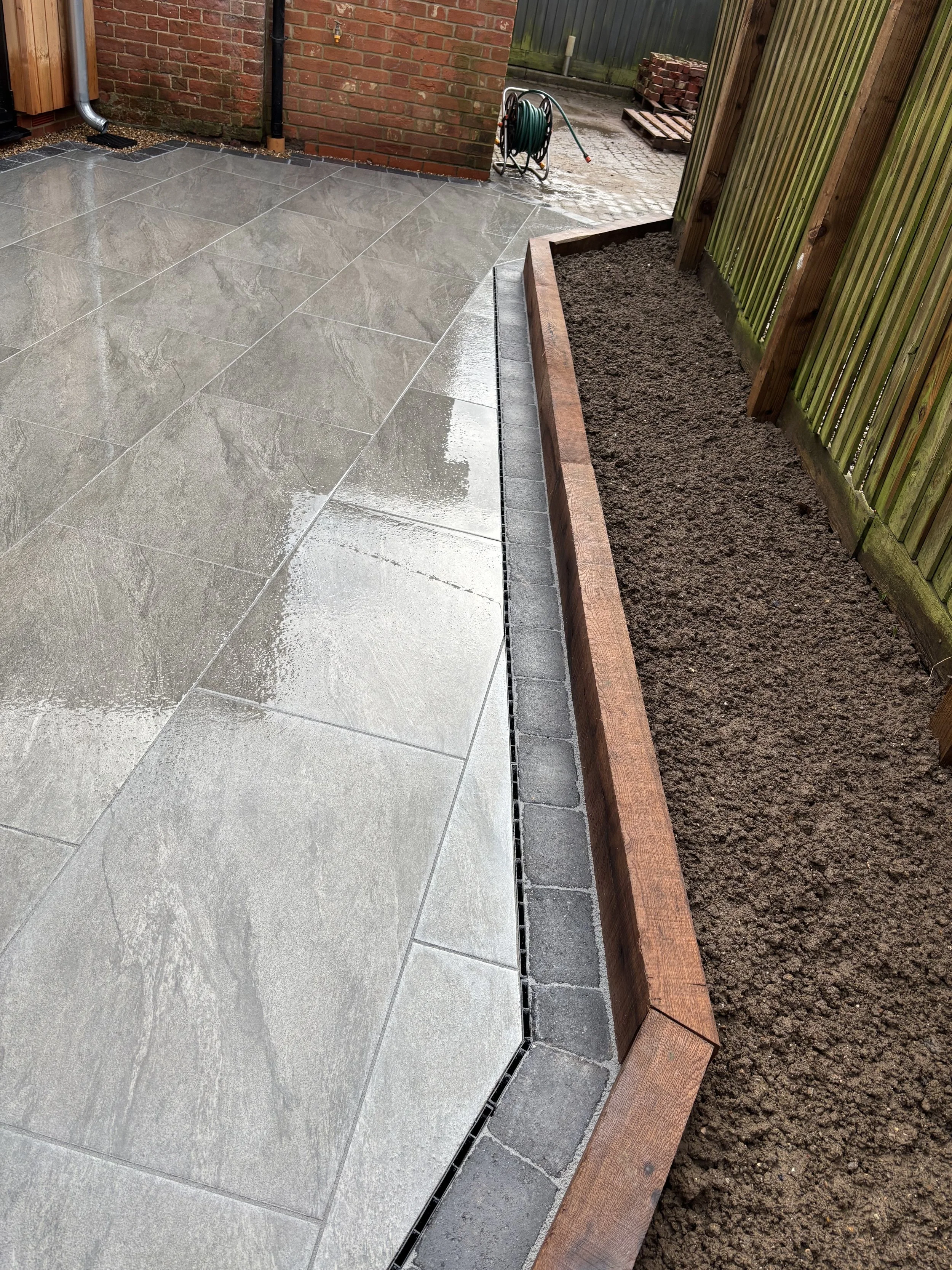 Wet gray tile patio area next to a garden bed with dark soil, bordered by a wooden plank and green fence, with a garden hose reel nearby.