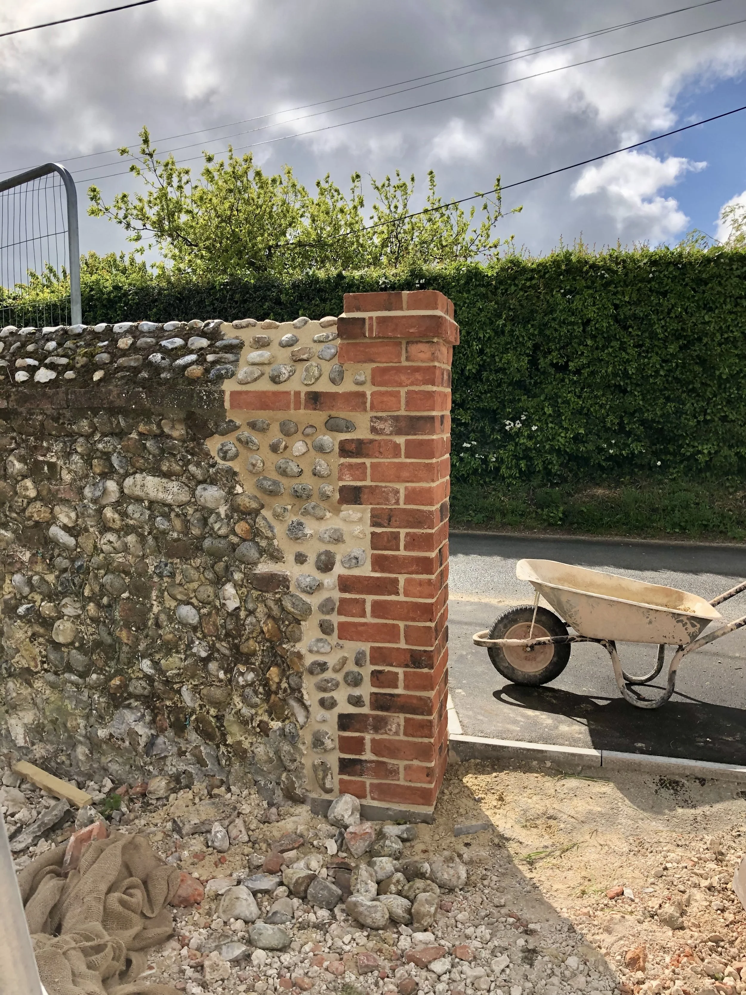 Partial brick and stone wall under construction, with a wheelbarrow nearby, and a parked wheelbarrow on a paved street, green hedges, trees, and a cloudy sky in the background.