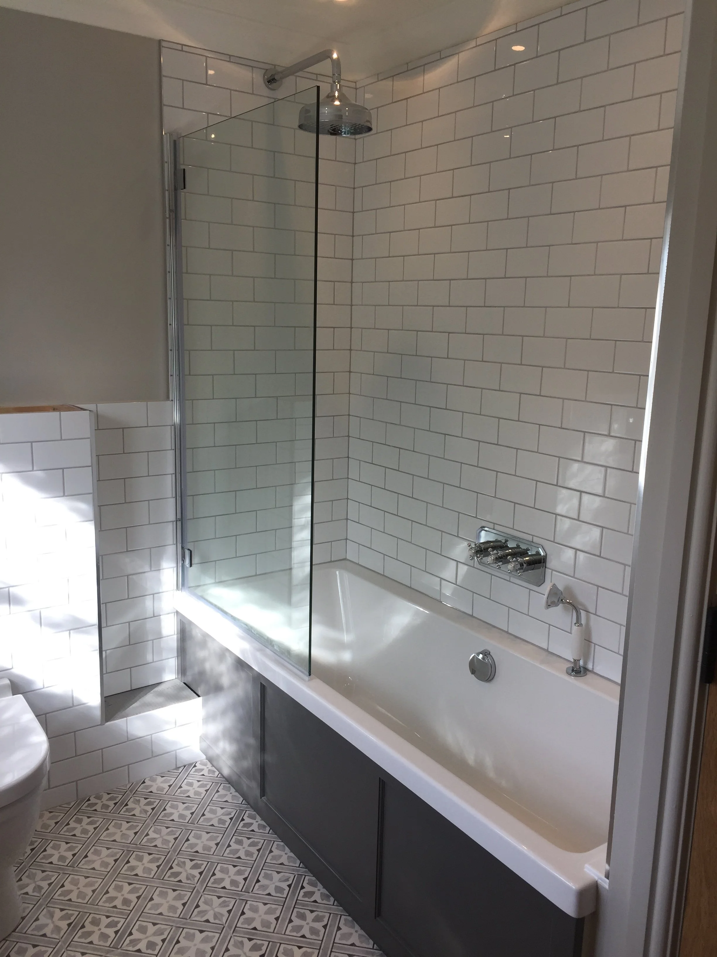 Bathroom with a bathtub, a glass shower partition, and white subway tile walls. The bathtub has a modern faucet, and the bathroom floor features patterned tiles.