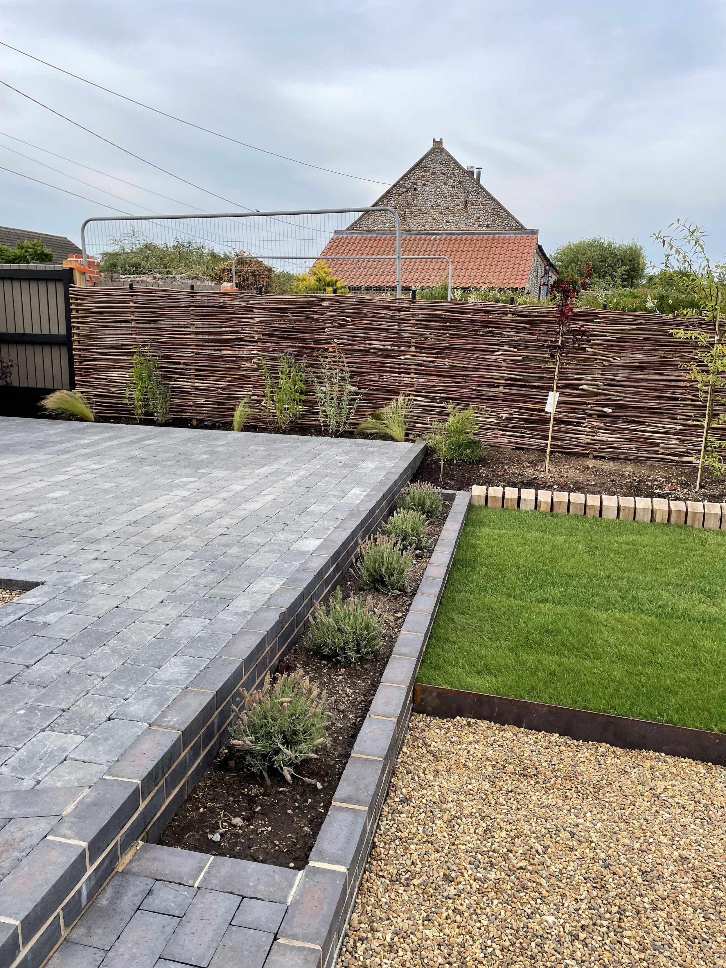 A backyard garden with a stone patio, a green lawn, a wooden fence, and a house with a red tiled roof and stone exterior under a cloudy sky.