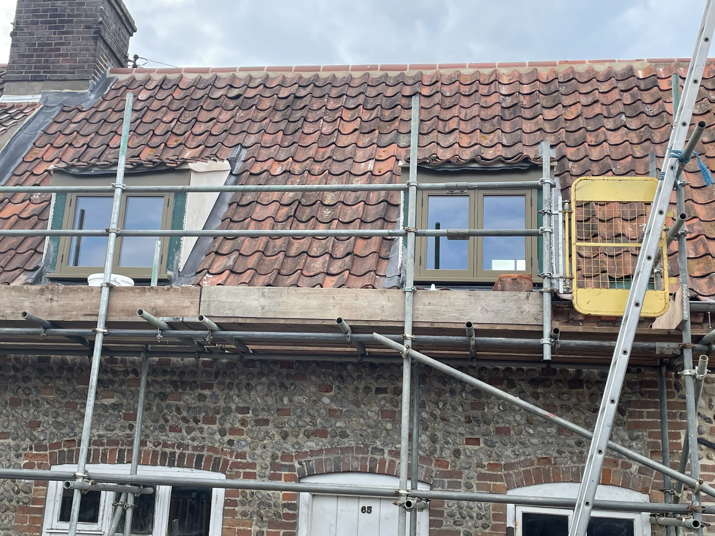 Roof of a building under construction with scaffolding, showing weathered red tiles, windows, and a brick chimney against a cloudy sky.