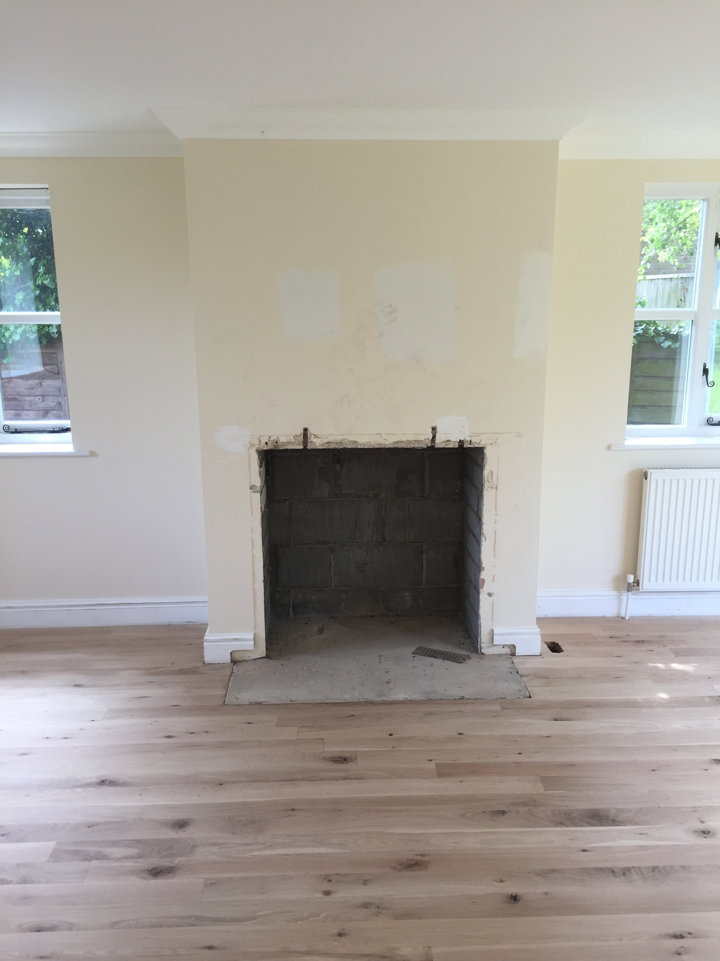 Living room with a fireplace under renovation, two windows, and light wood flooring.