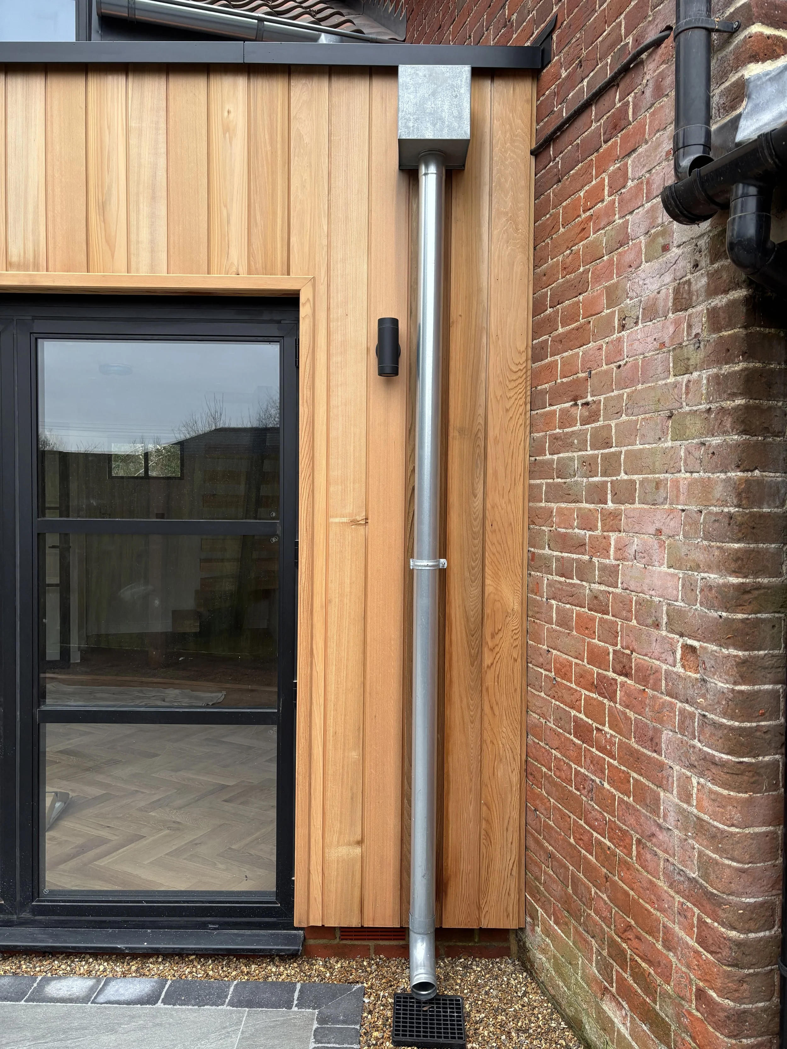 Metal drainage pipe attached to a wooden wall beside a brick wall, with a small black drainage grate at the bottom.