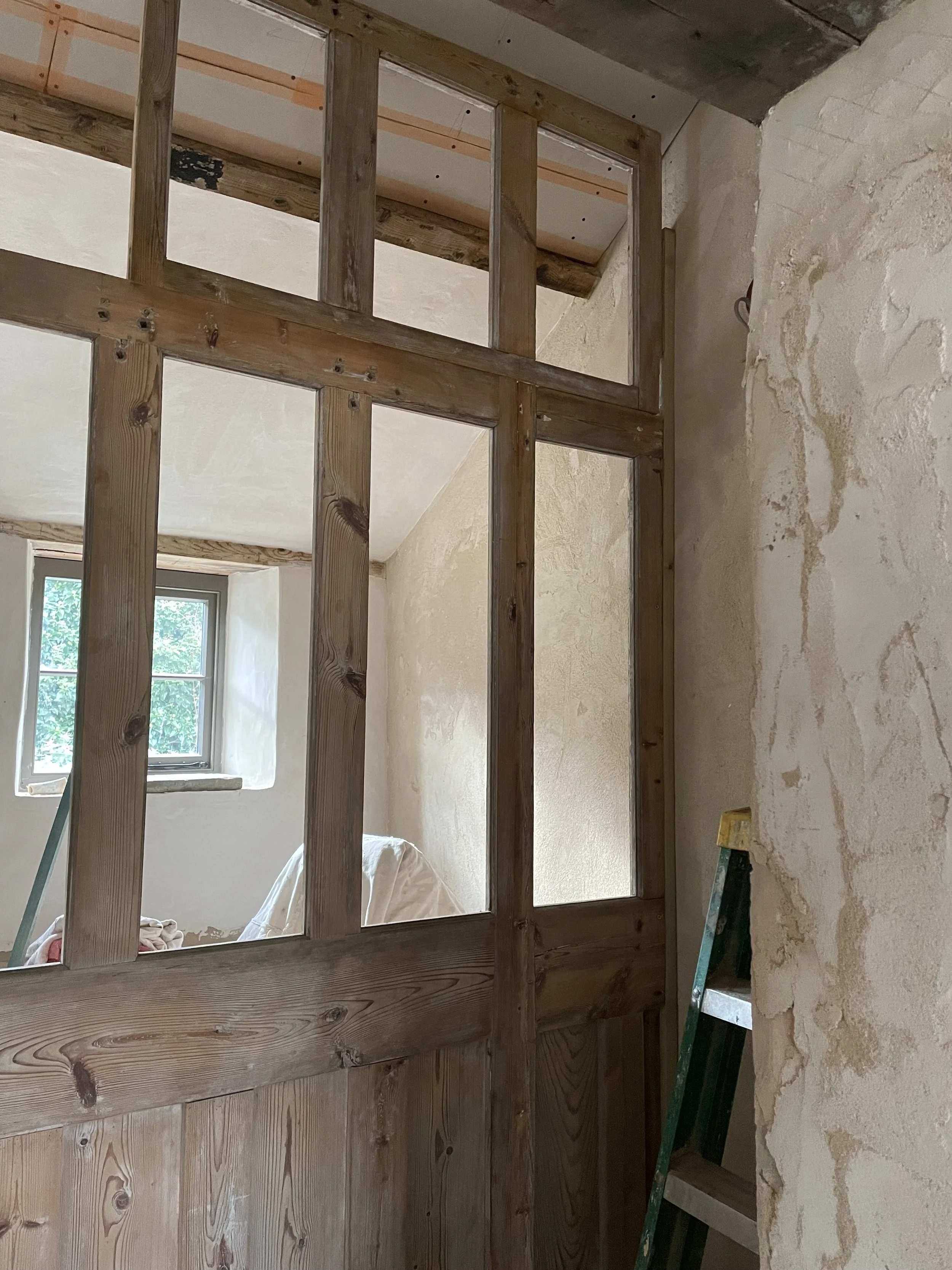 Interior of a room under renovation with wooden framing and a small window, walls partially plastered, a ladder leaning against the wall.