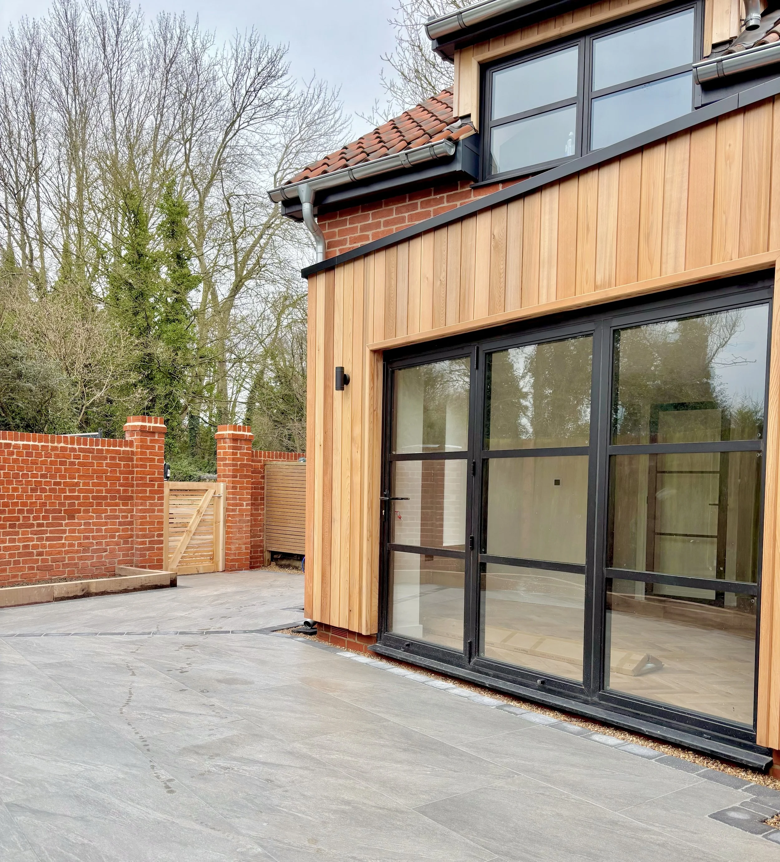 Part of a modern house with large glass doors, wooden siding, and a brick section near the roof, with an outdoor patio area and a brick wall in the background, surrounded by leafless trees.