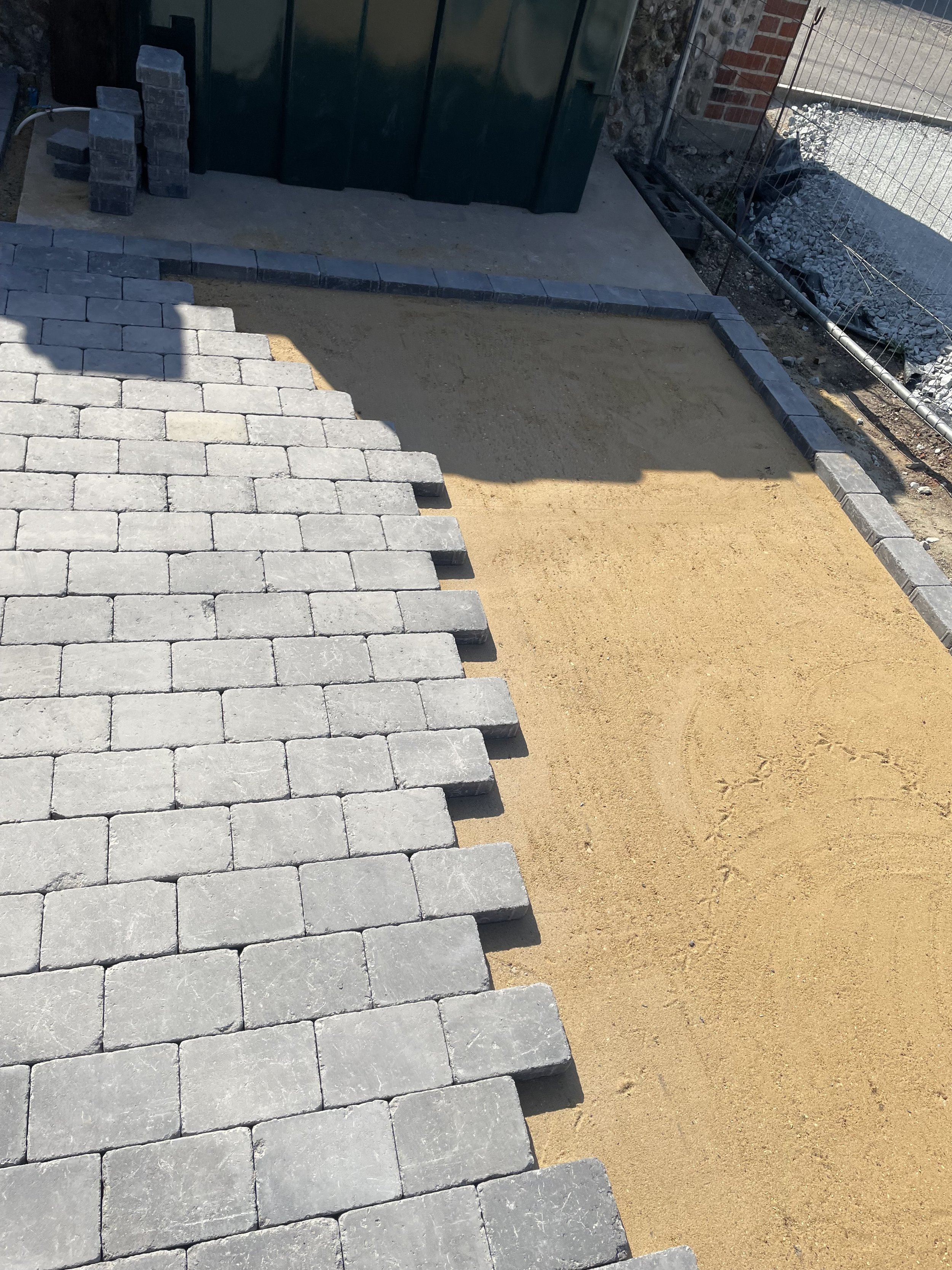 Part of a patio or walkway under construction with gray paving stones partially laid, a pile of unlaid yellow sand, and a small area of bare concrete. Background shows a green metal shed, a brick wall, and a metal fence.
