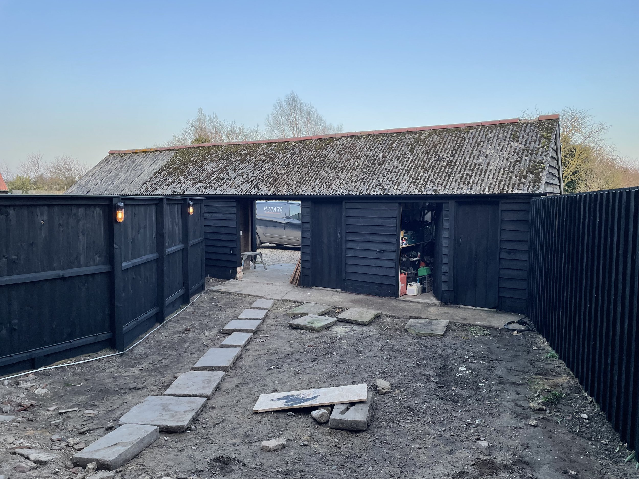 A backyard scene showing a black wooden shed, a row of stepping stones on dirt, and a fence with black panels. There is a garage with an open door, some tools inside, and a parked van seen through the opening. The roof of the shed appears to be weath