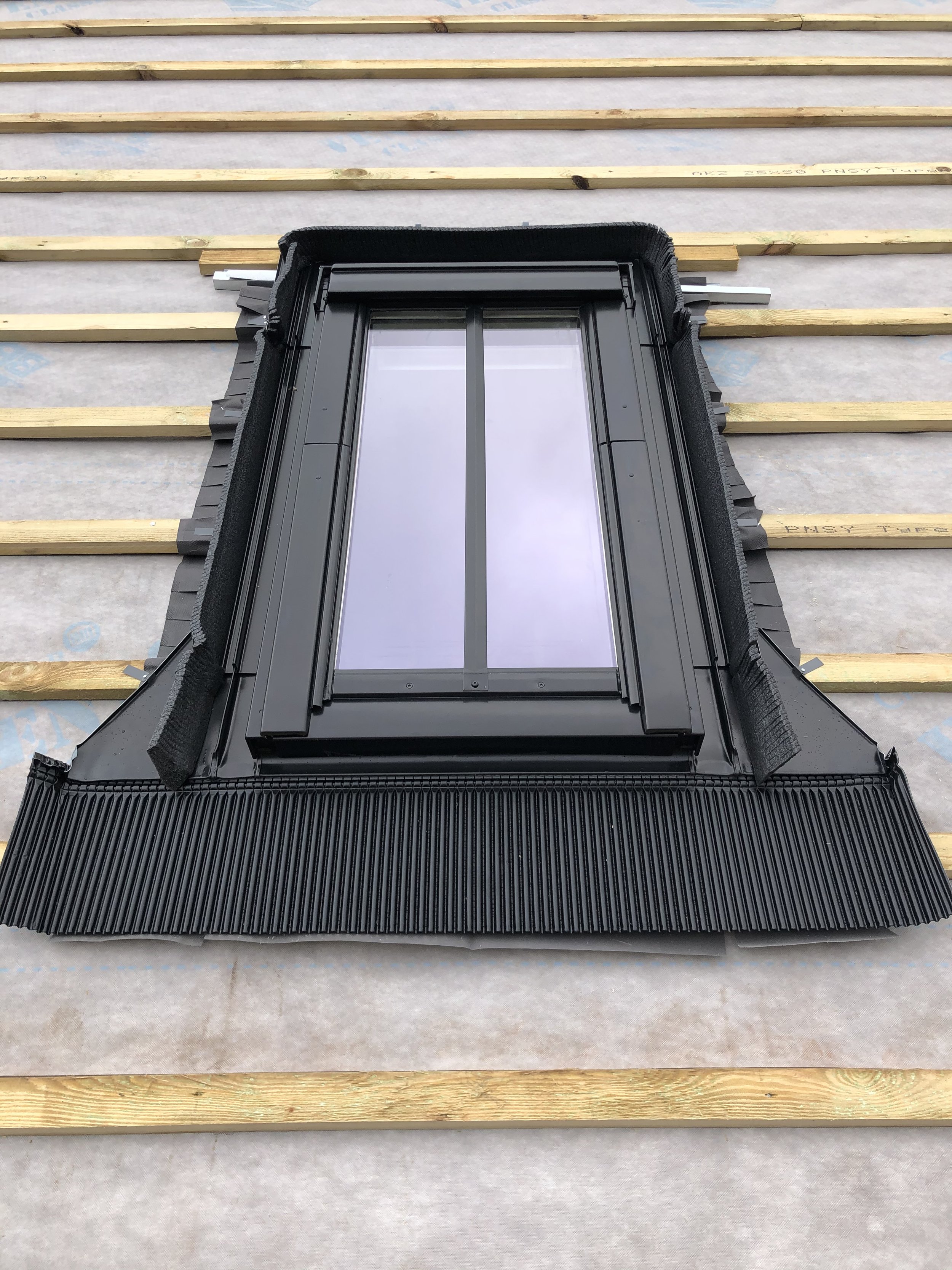 A black roof window installed on a roof under construction with wooden battens and roofing paper visible.