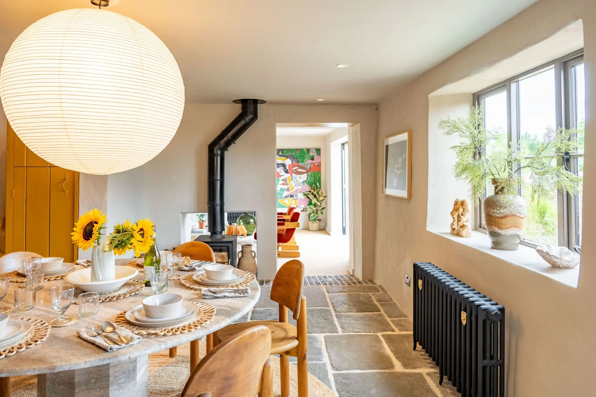 A cozy dining room with a round table set for a meal, surrounded by wooden chairs. The table features sunflowers in a white vase, with plates, glasses, and silverware arranged. Large windows on the right let in natural light, and a black radiator is 