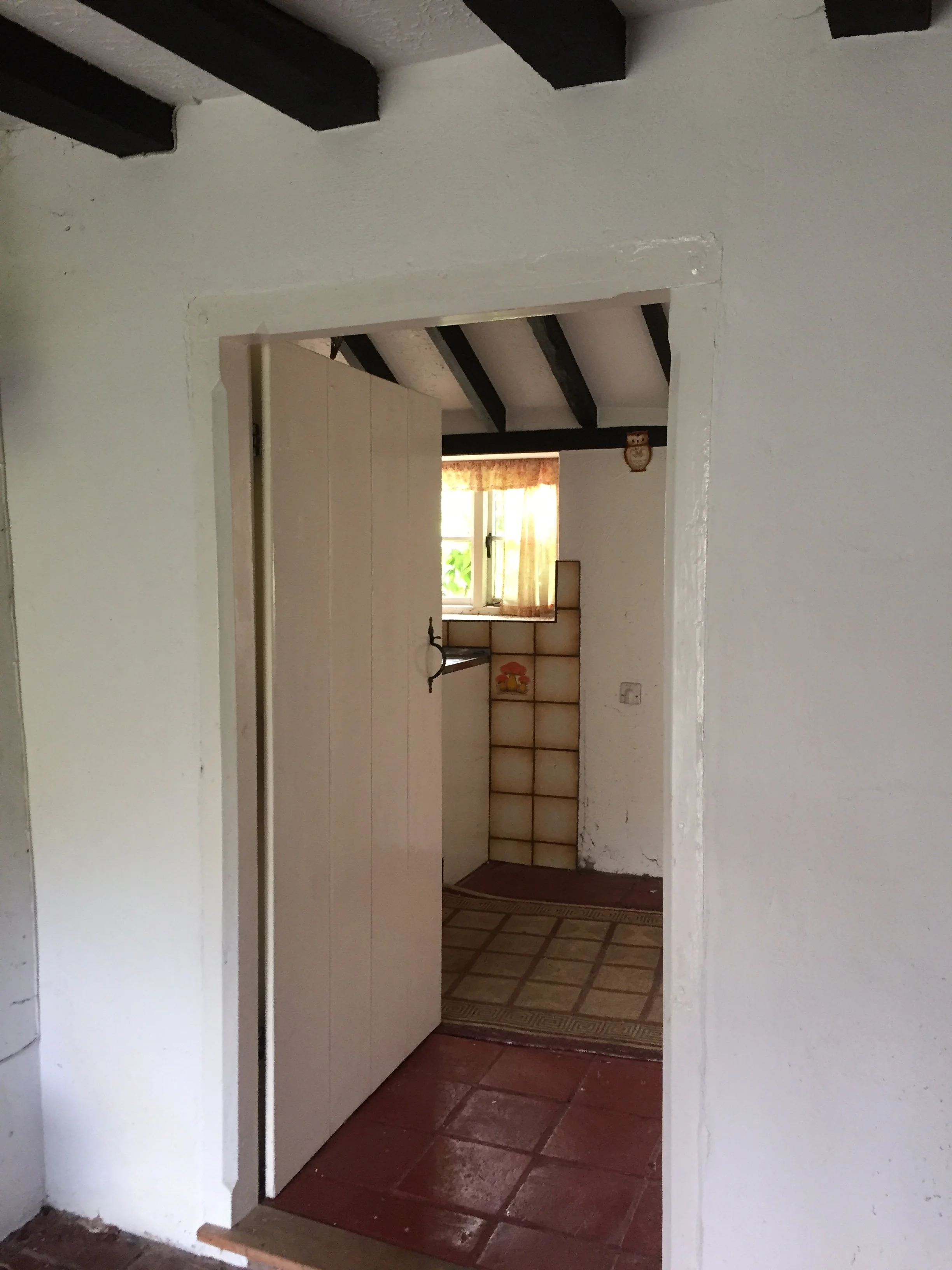 Interior view of a house hallway with a doorway leading to a room. The room has a window with curtains, a small piece of furniture, and a wall decoration of an owl. The hallway has tile flooring and white walls with visible texture.