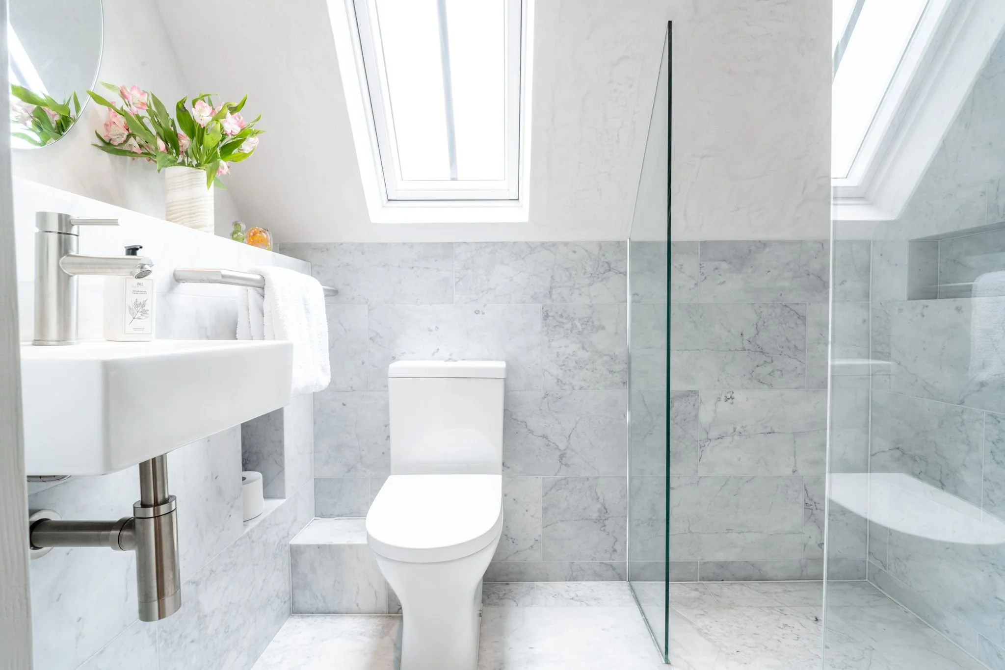 A modern bathroom with white marble walls and a skylight window. There is a white sink with a soap dispenser and a towel hanging on a rail. A toilet is visible beneath the window, and on the ledge above the toilet, there is a flower vase with pink an