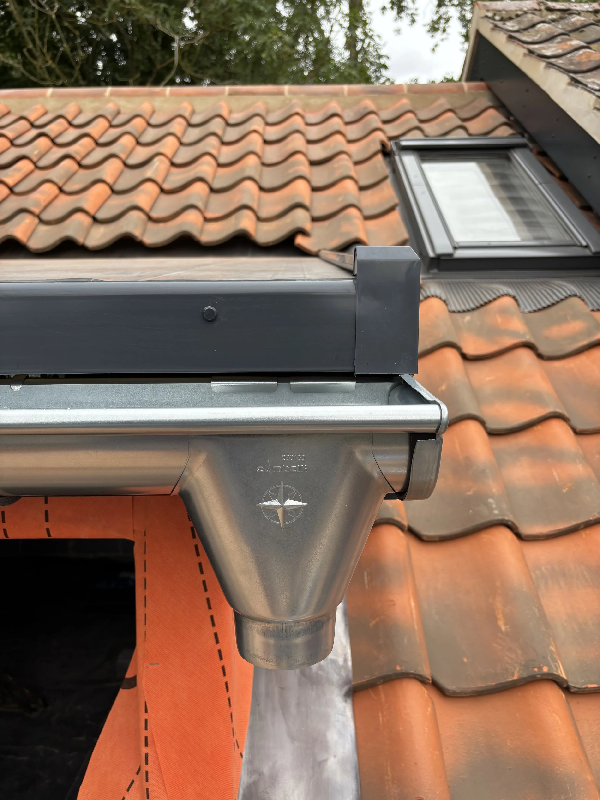 Close-up of a metal chimney flashing installed on a tiled roof with a skylight window.