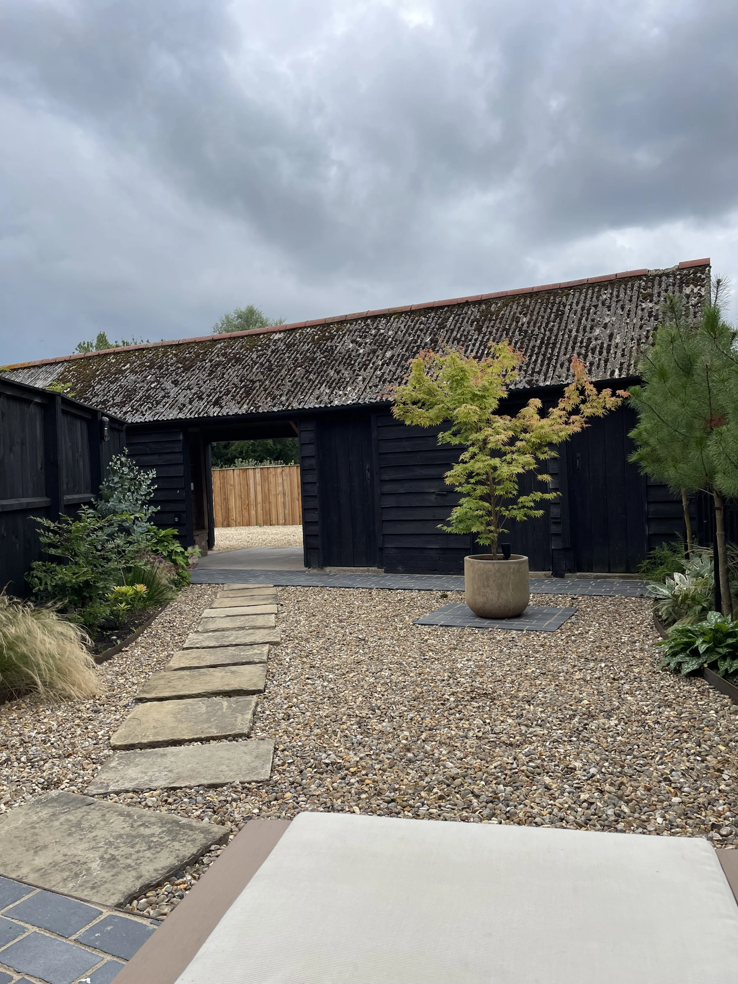 A backyard garden with a stone pathway, a gravel yard, potted plants, a black wooden shed, and trees, under a cloudy sky.