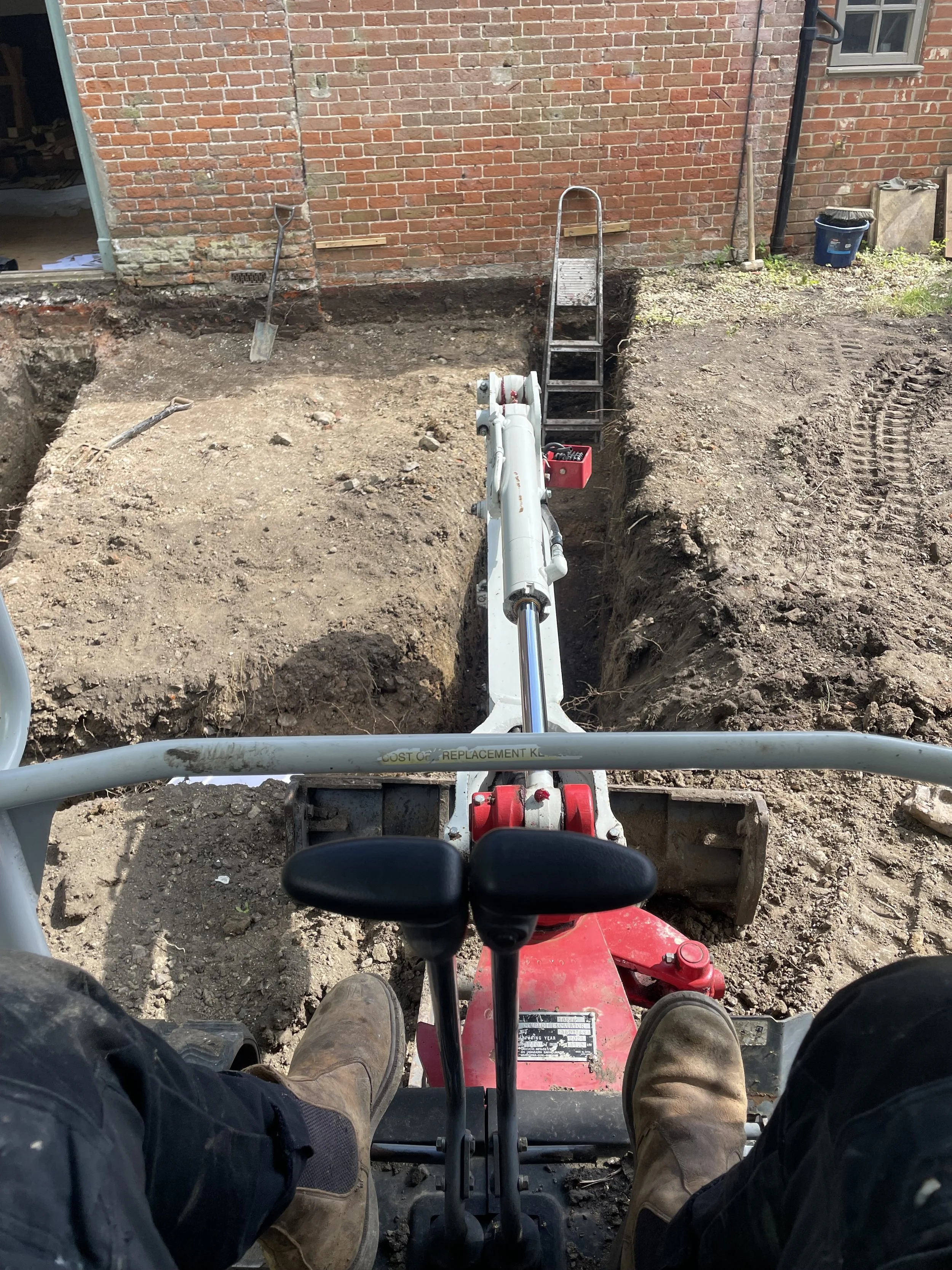 First-person view of a person operating a mini excavator, with their legs and boots visible, digging a trench in a backyard next to a brick house. The excavator's arm and bucket are in the foreground, and a ladder and tools are visible in the backgro