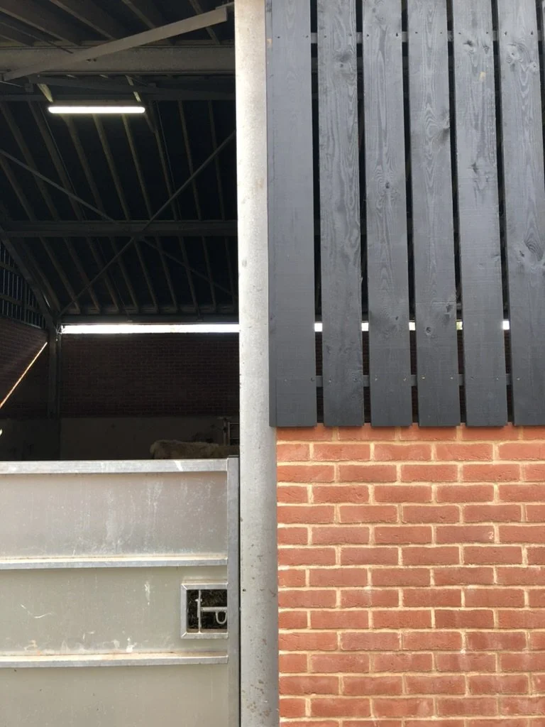 A close-up of the exterior corner of a building showing a section of grey metal fencing, a brick wall, and a metal grain silo or storage container.