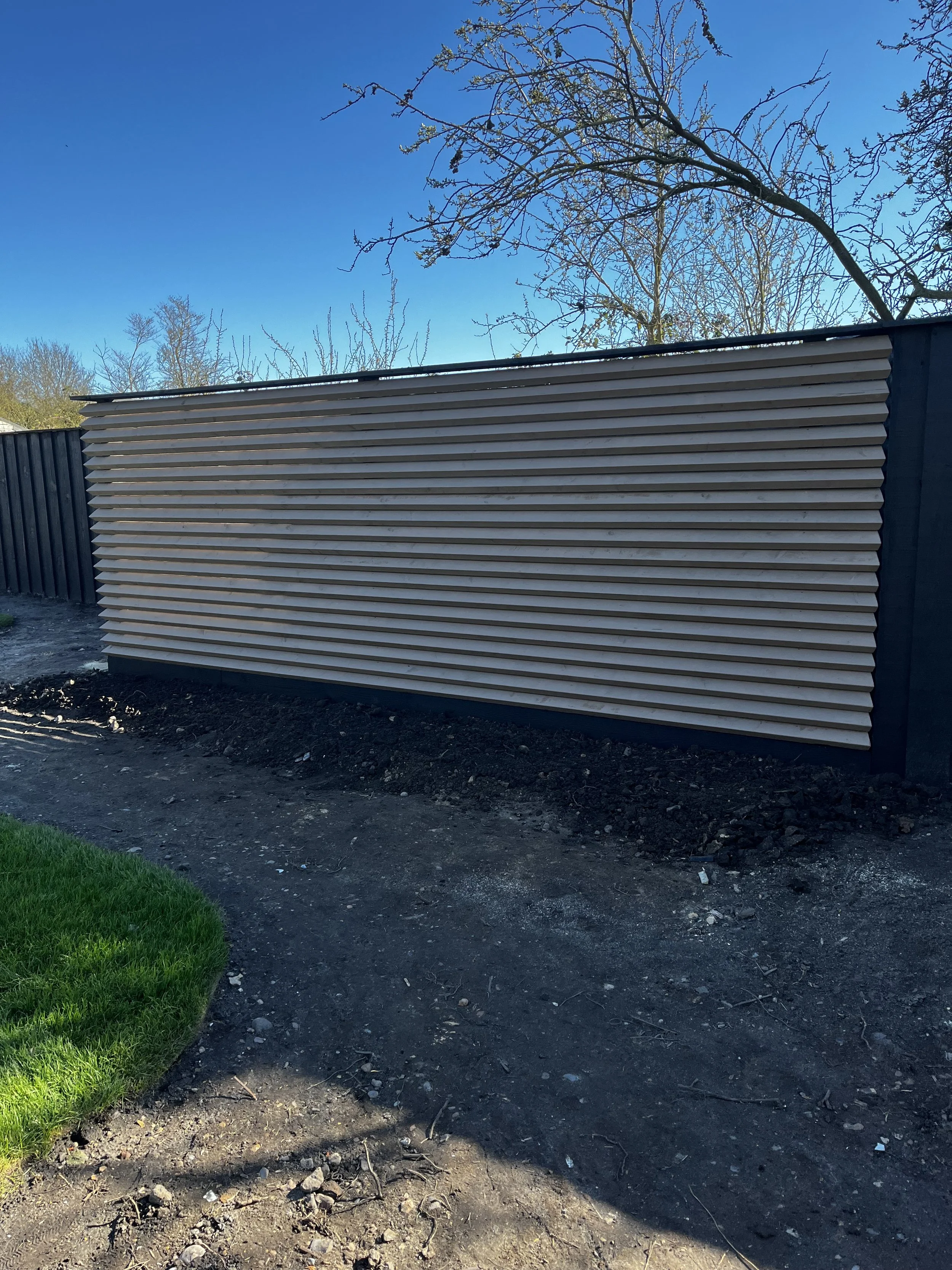 A newly installed wooden fence with horizontal slats, with a background of blue sky and leafless trees. The ground in front is dirt and some patches of green grass.