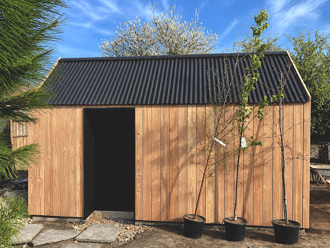 A wooden shed with vertical planks and a black corrugated metal roof, three potted trees in front, and a blue sky with clouds overhead.