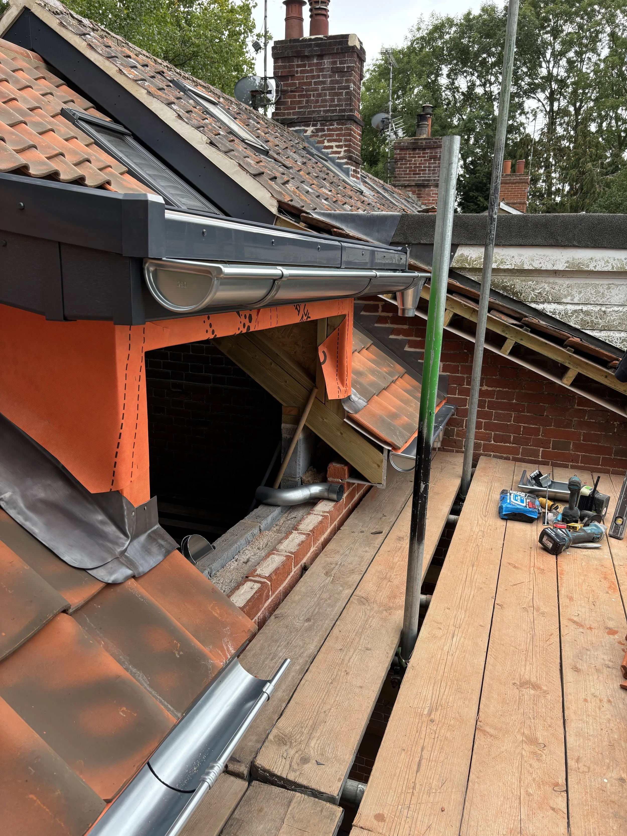 A sloped brick roof with new roofing materials being installed, including a metal gutter, scaffolding, and tools on a wooden platform.