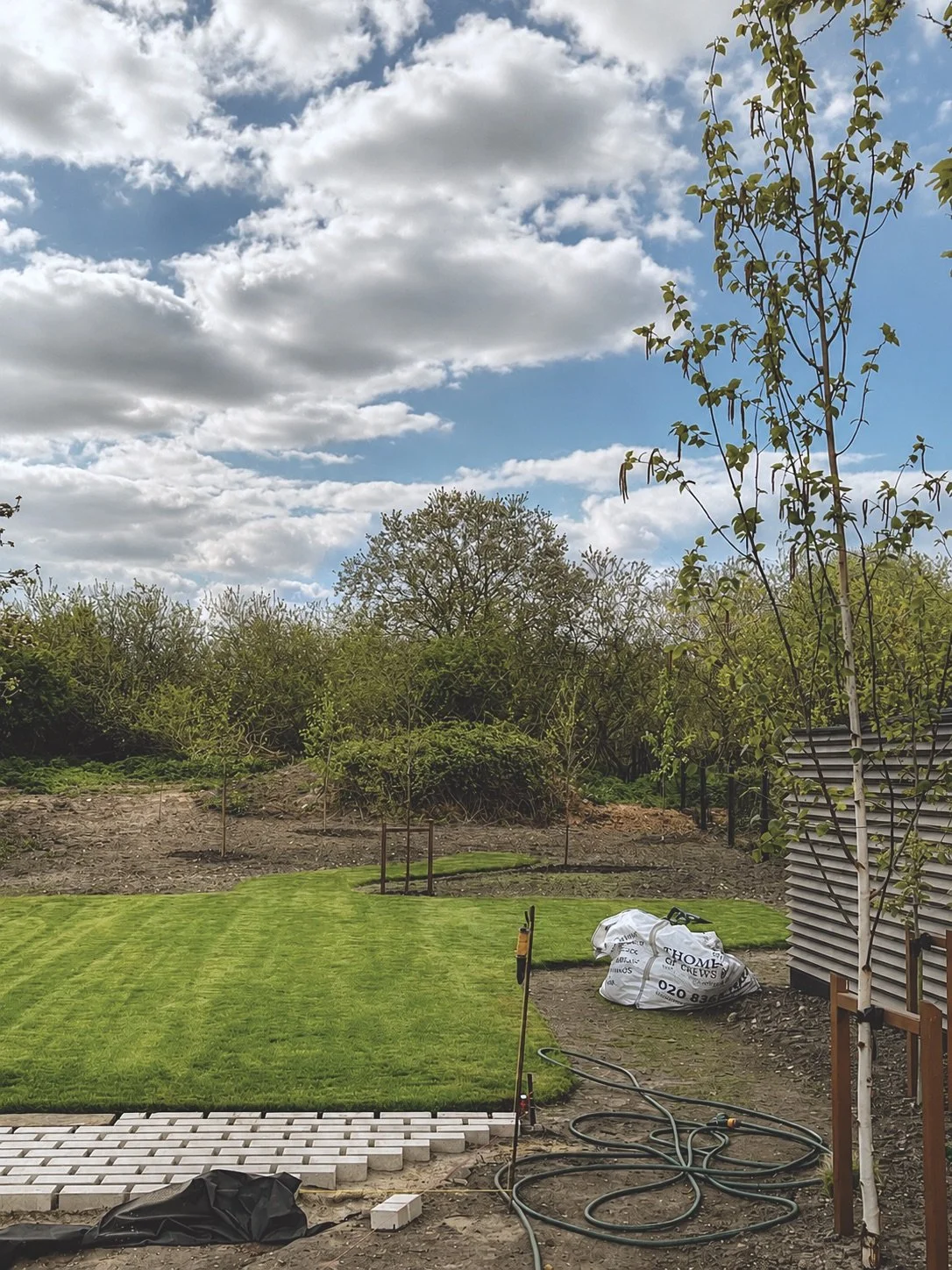 A backyard garden during daytime with freshly mowed grass, young trees, and construction supplies including a bag of materials and hoses near a building with modern siding.