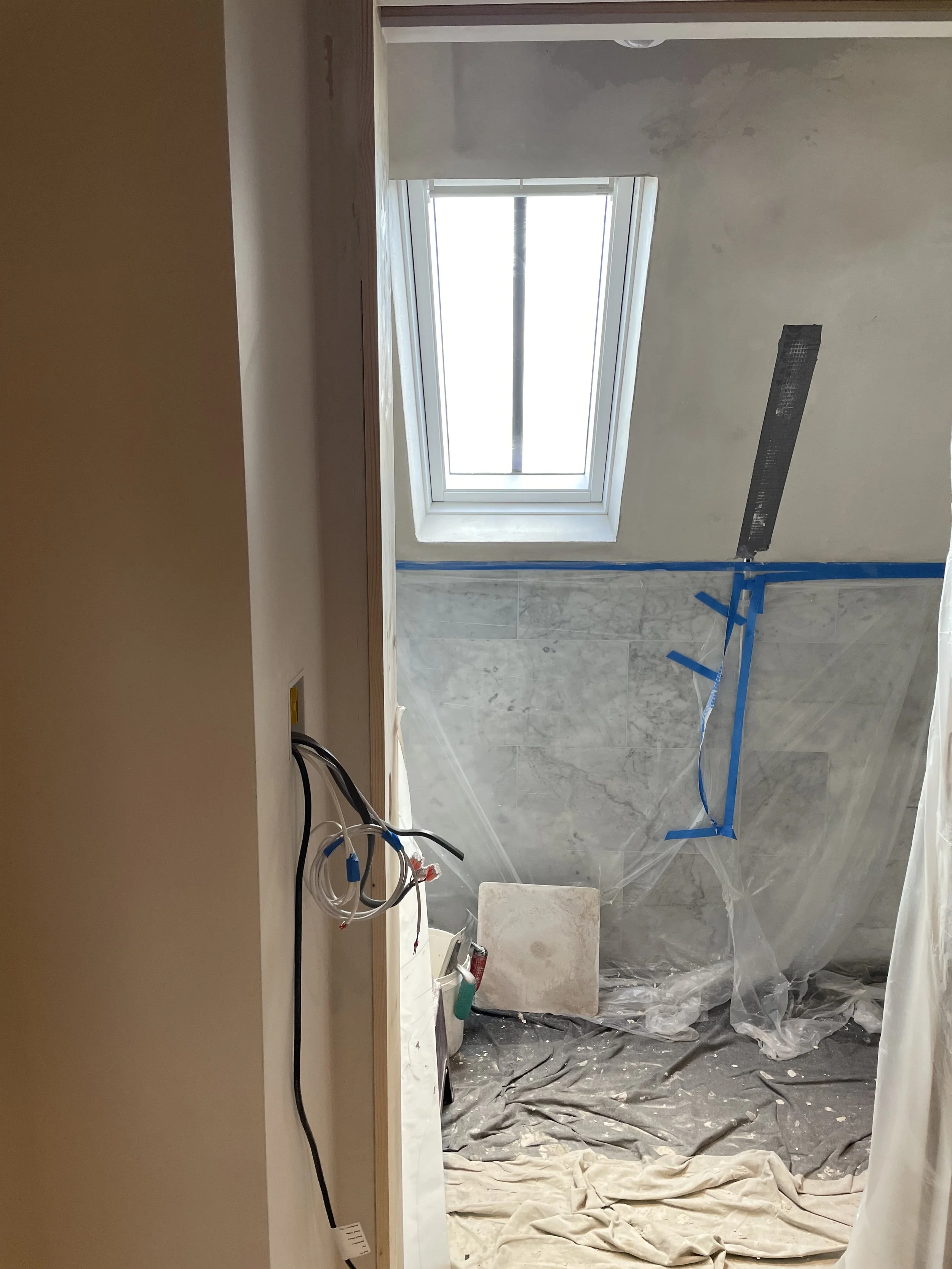 A room under renovation with a window on the back wall, construction tools, cords hanging from the wall, plastic sheets on the floor, and some drywall and construction materials around.