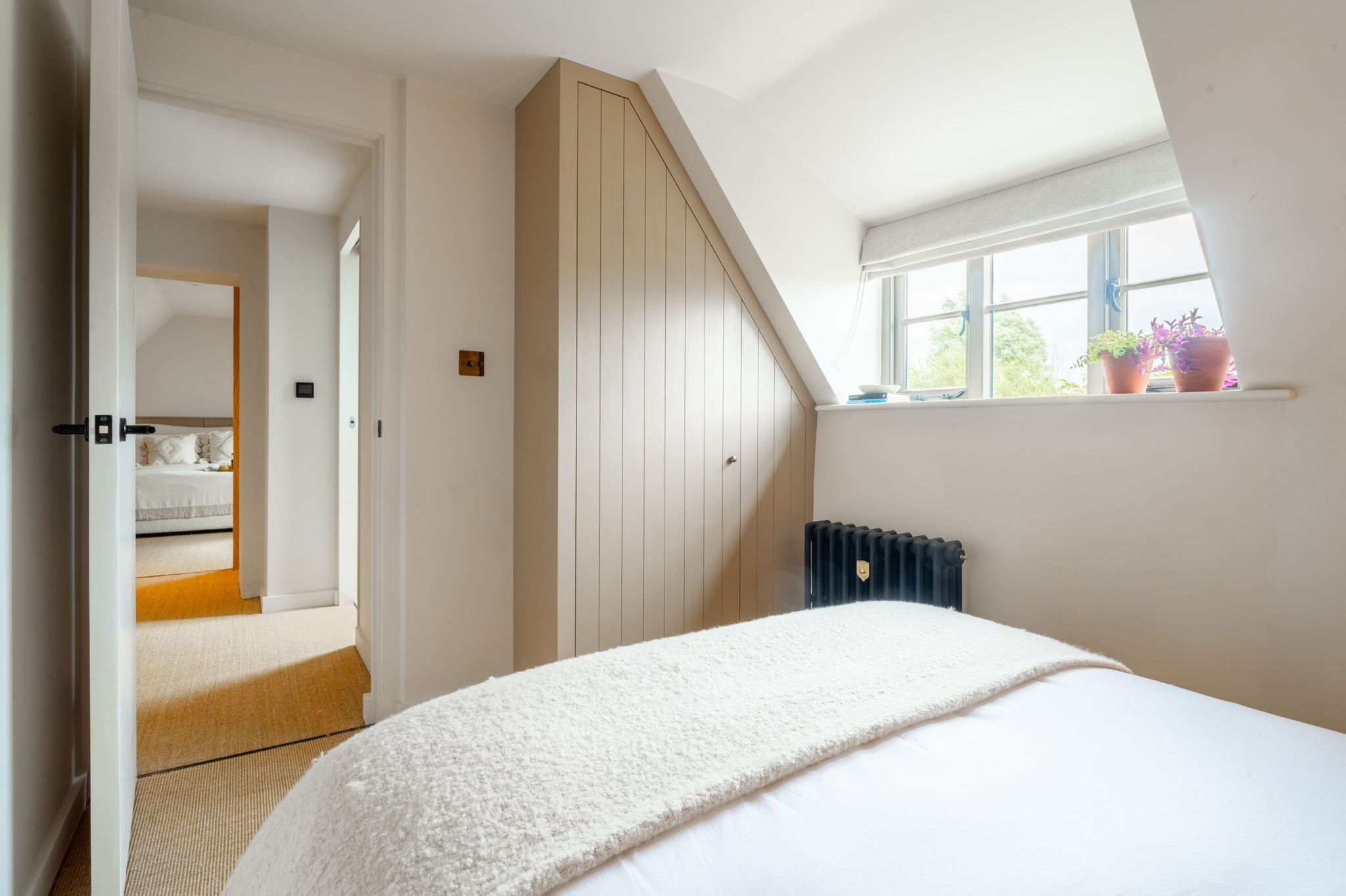 A cozy bedroom with a white bedspread, a window with potted plants, and a view of a tree outside. An open door leads to a small hallway with a bedroom visible through the doorway.