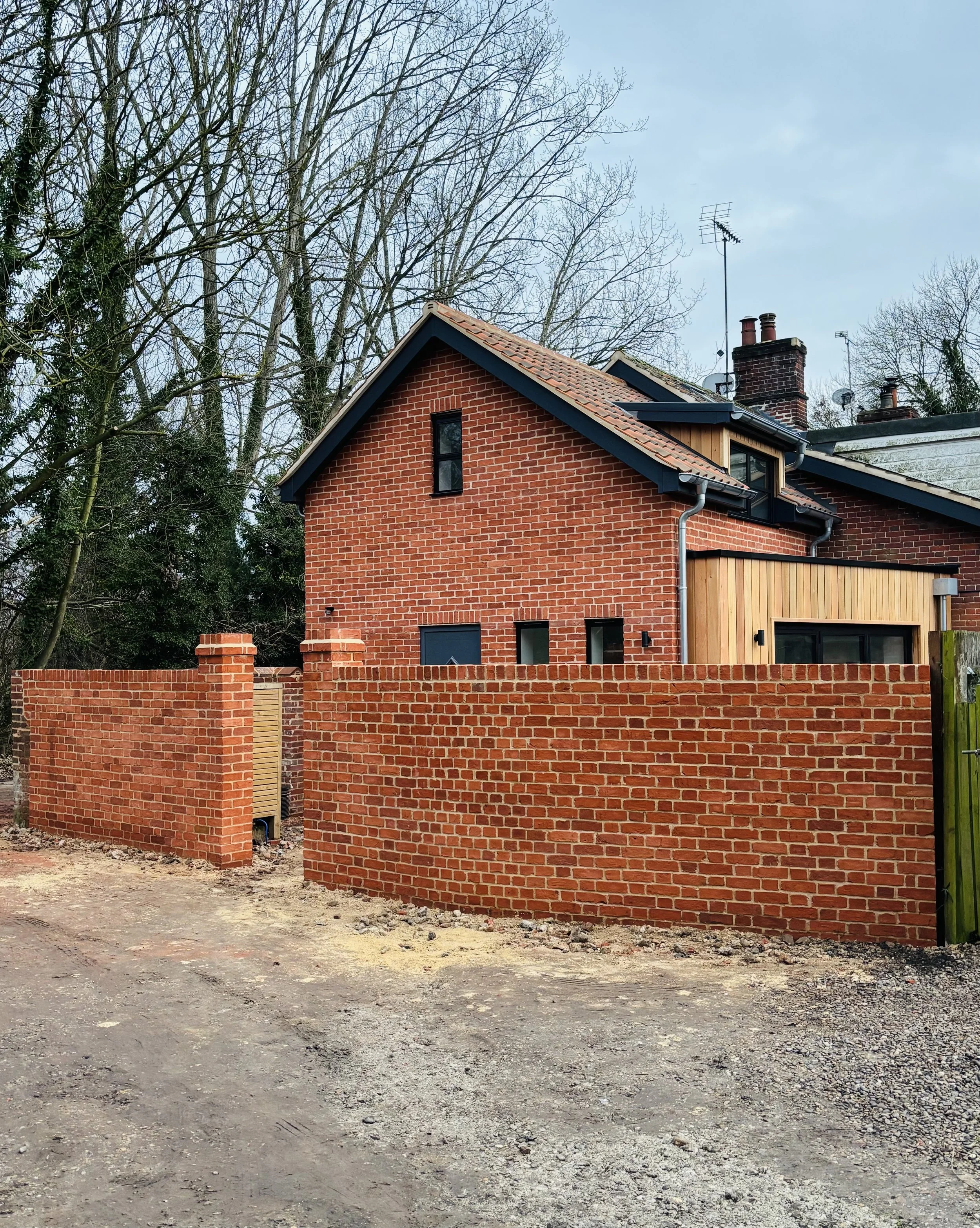 A red brick house with a partially completed brick fence in front of it, set against a background of leafless trees and a cloudy sky.