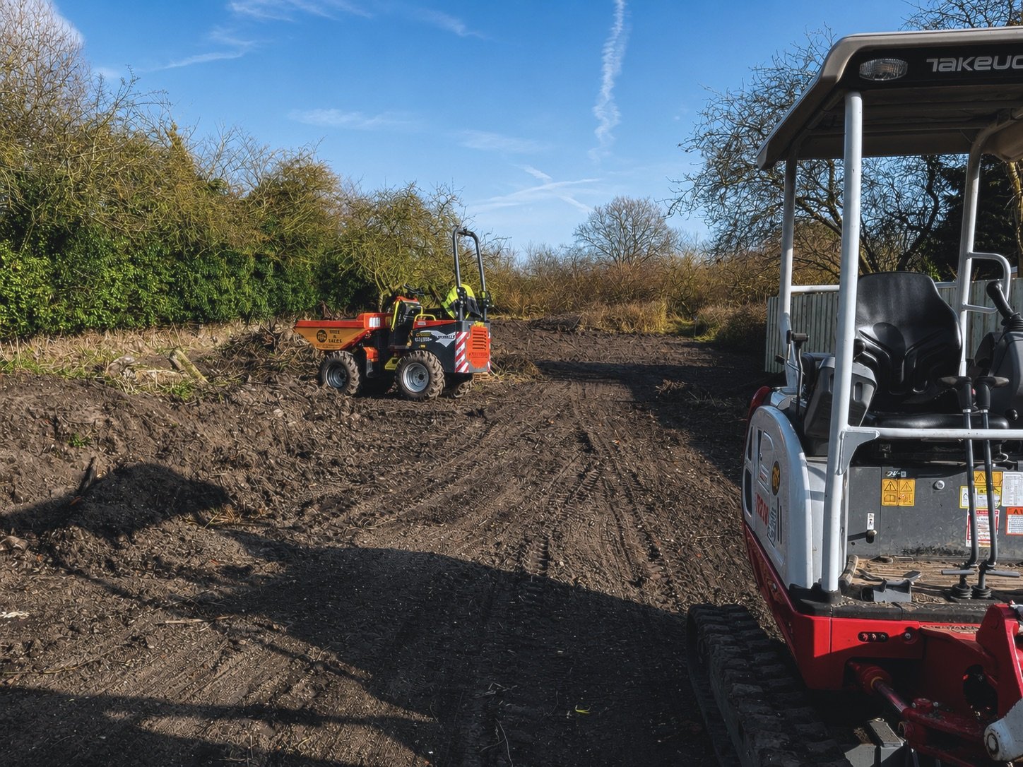 Construction site with small bulldozer and excavator on dirt ground, surrounded by leafless trees and bushes against a blue sky.