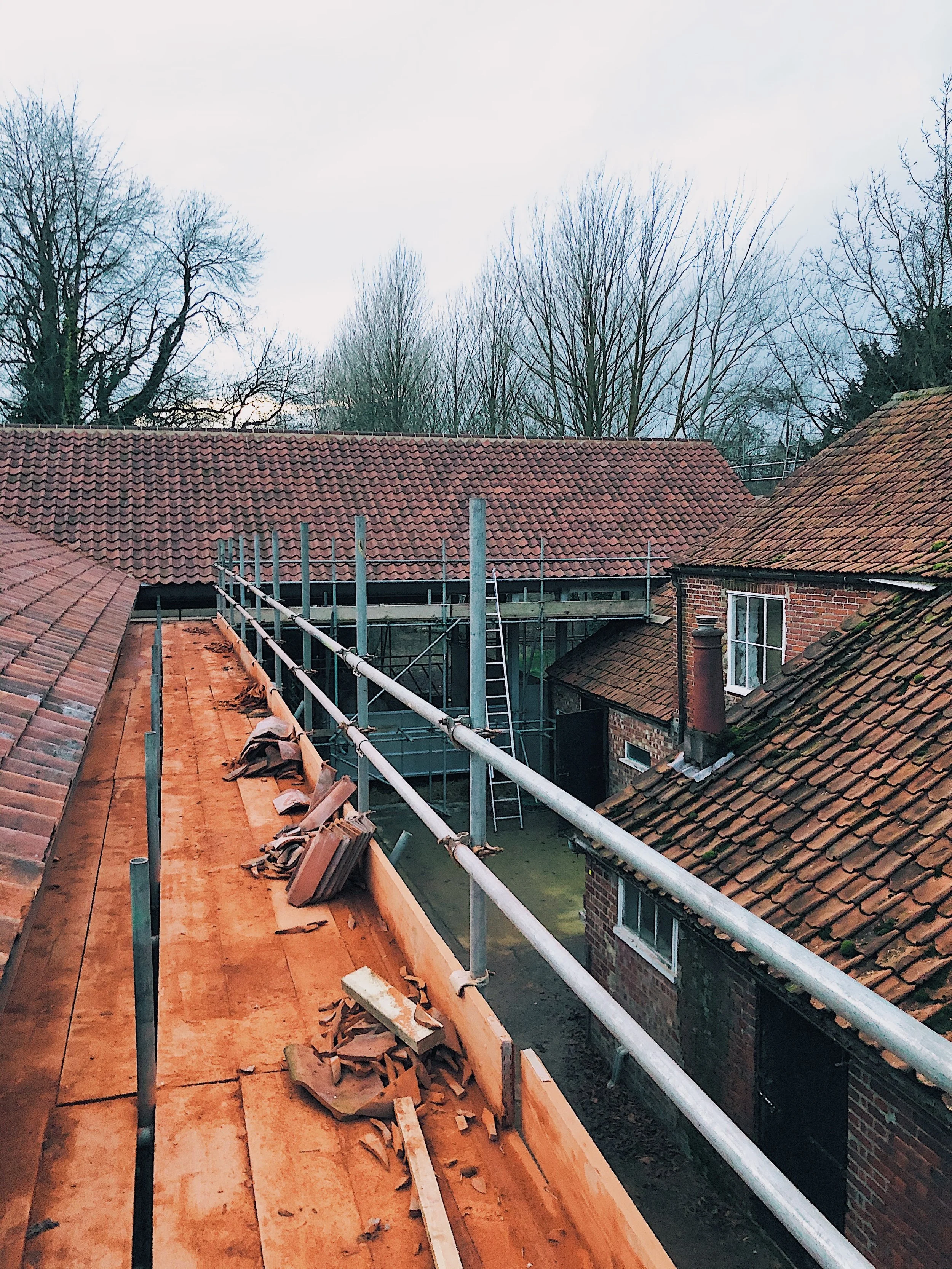 View from a rooftop under construction, showing scaffoldings and rooftops of neighboring houses, with leafless trees and cloudy sky in the background.