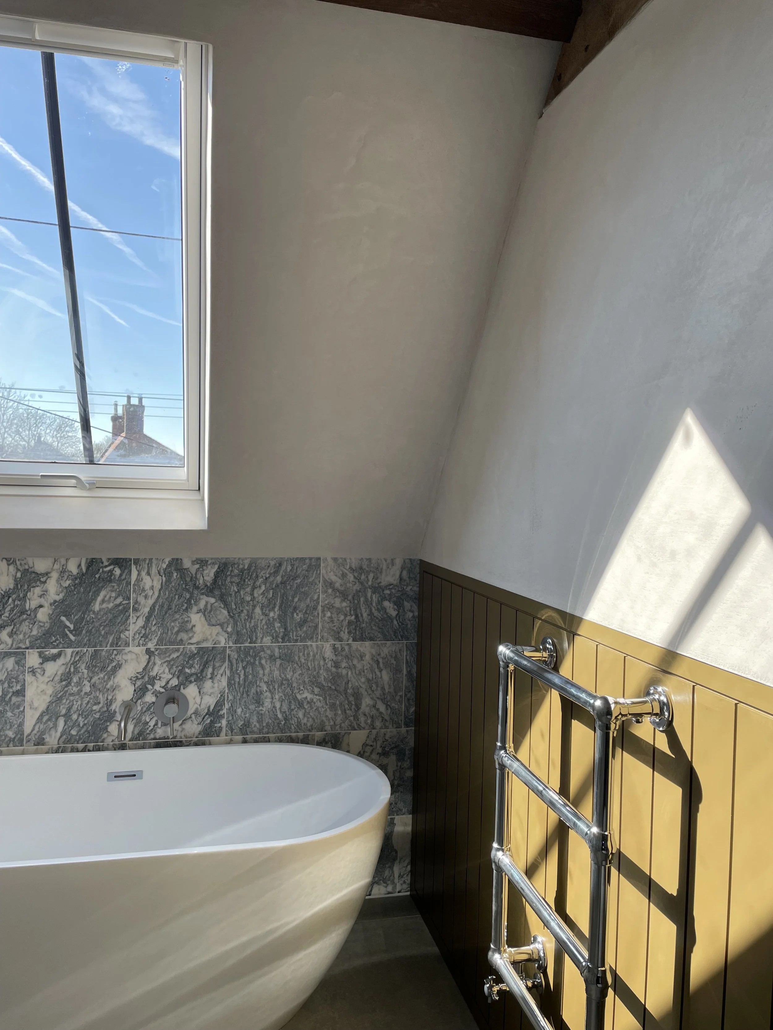 A bathroom with a white bathtub, black and white marble wall tiles, a window showing a blue sky with clouds and contrails, a wooden panel with a towel rack, and a shower faucet.