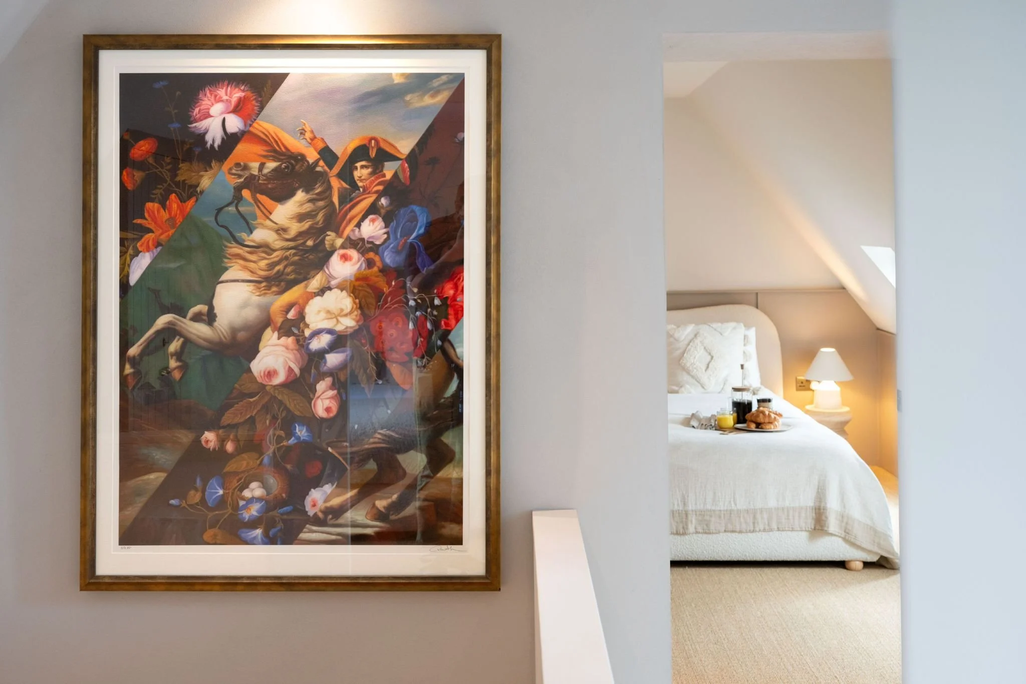 Framed colorful fantasy art of a woman with a bird hat, flowers, and animals, viewed from a bedroom with a white bed and nightstand.