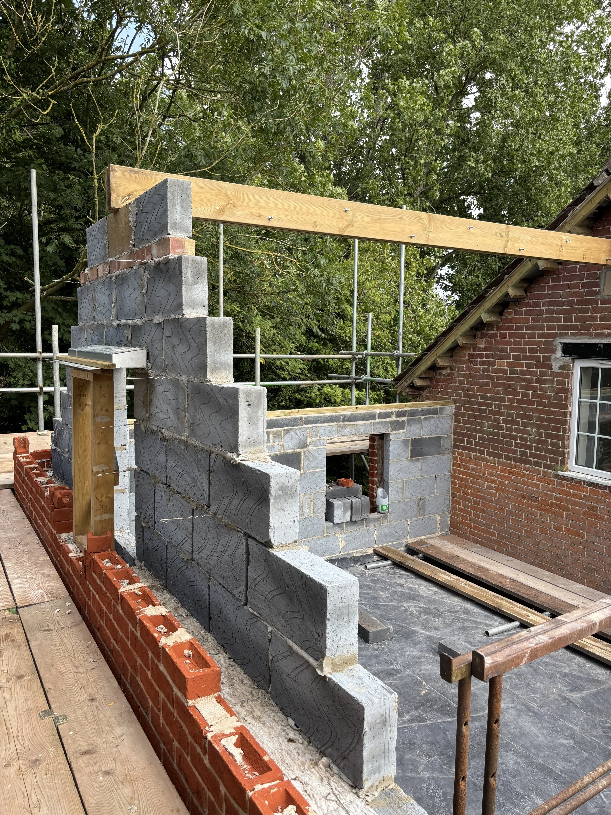 Construction site with partially built brick and concrete wall, wooden beams, and scaffolding, next to a brick house with a window and background trees.