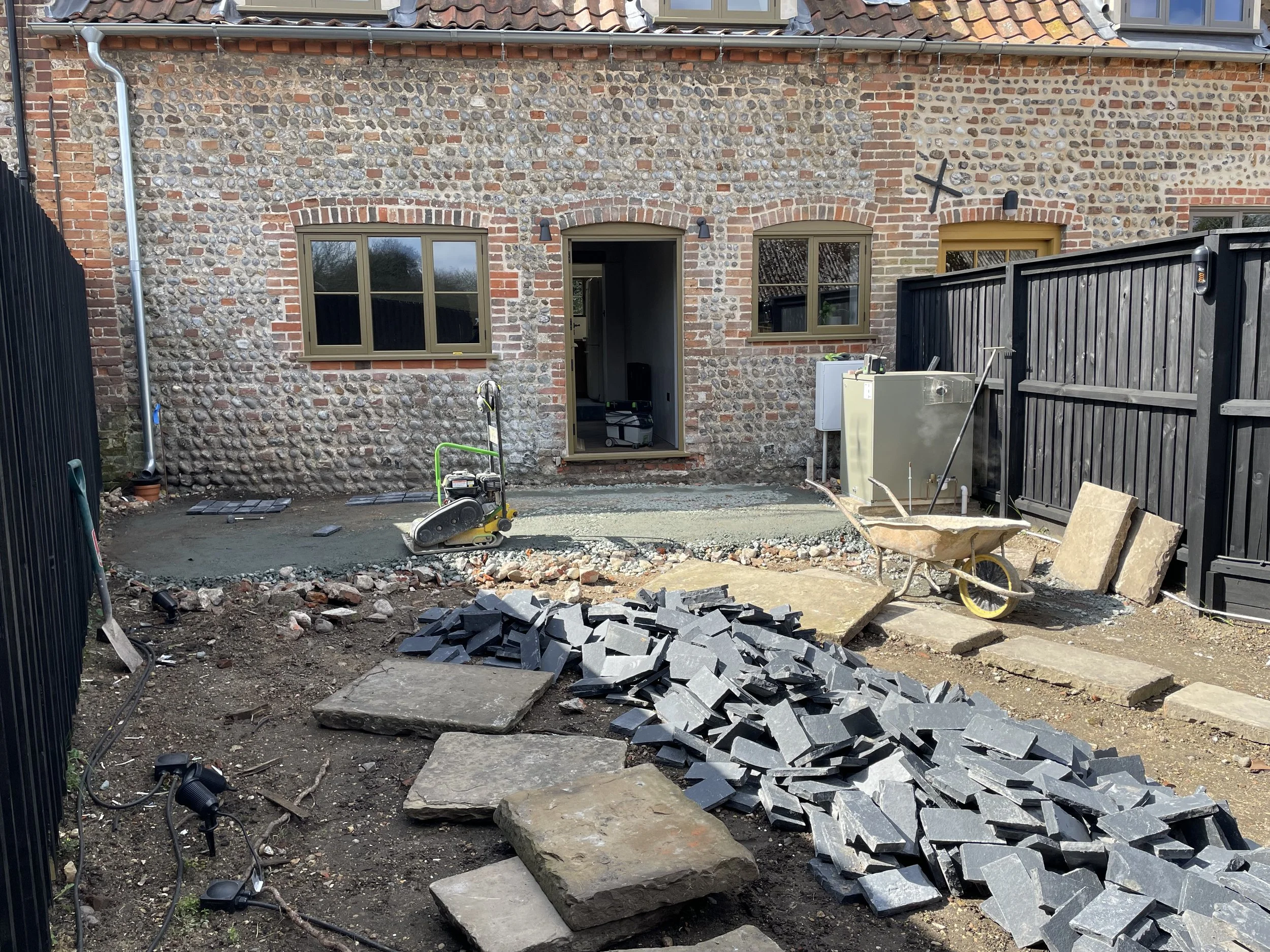 Backyard under renovation with construction tools, a pile of gray paving stones, a wheelbarrow, and construction materials. The house has brick and stone exterior walls with three windows and an open door. Fencing surrounds the area.