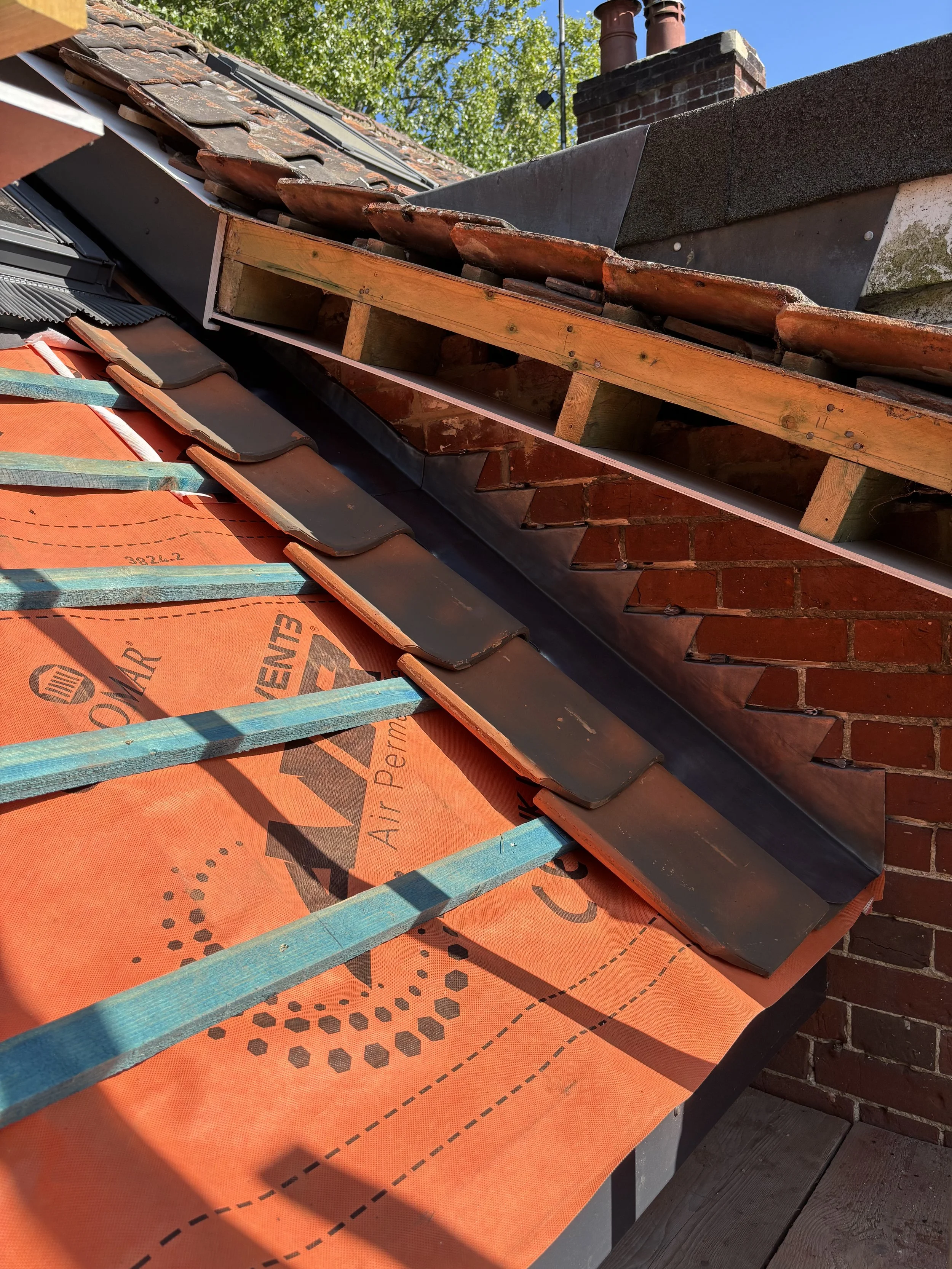 Close-up of a rooftop under construction showing orange roofing underlayment, blue wooden battens, black tiles, and brick chimney with chimney caps, with trees and chimney stacks in the background.