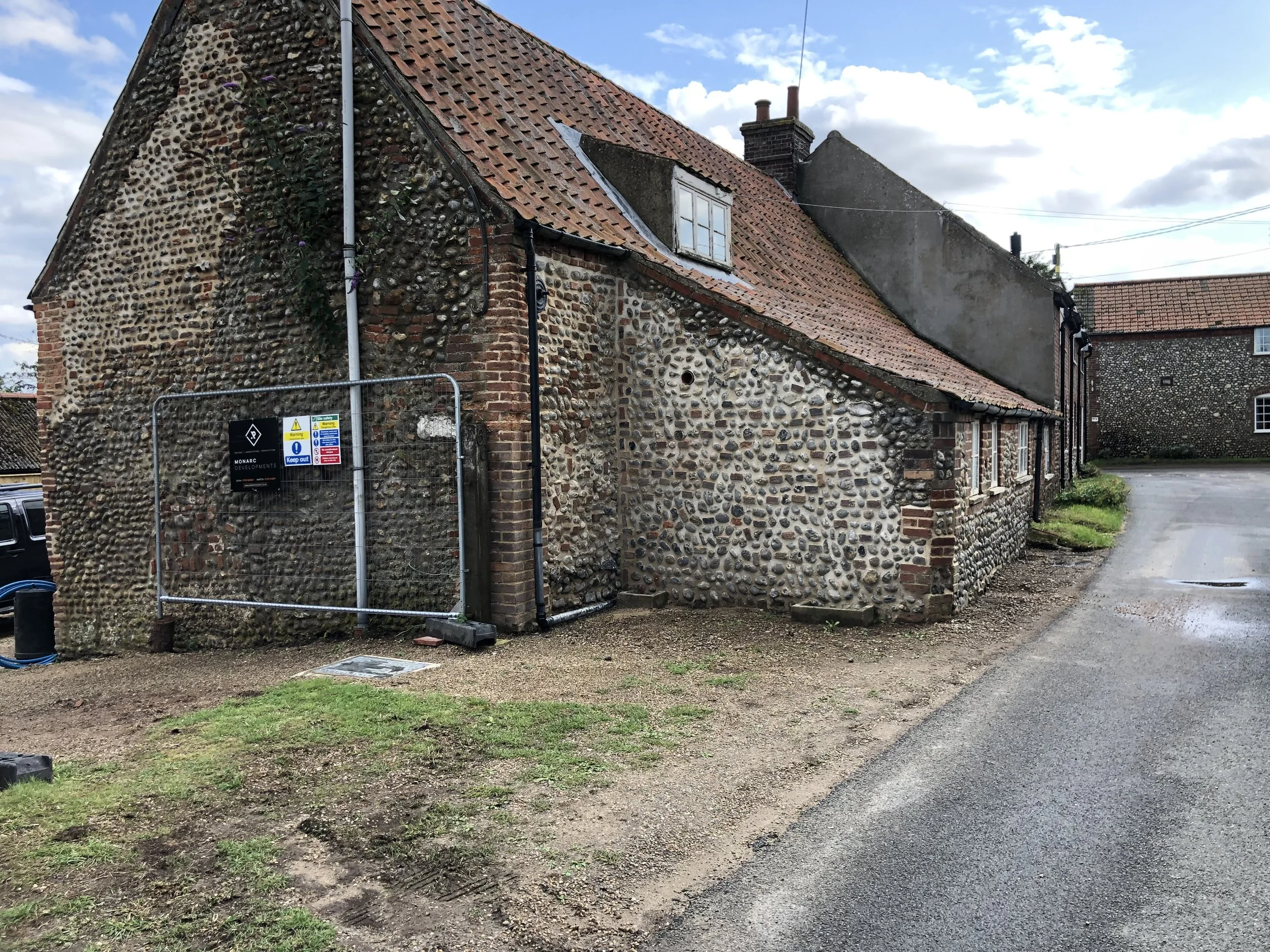 An old building with a brick and pebble exterior wall, a sloped red tile roof, and a small dormer window. The building has a chimney and appears to be in a rural or semi-rural area. There is a construction fence with warning signs at the corner of th