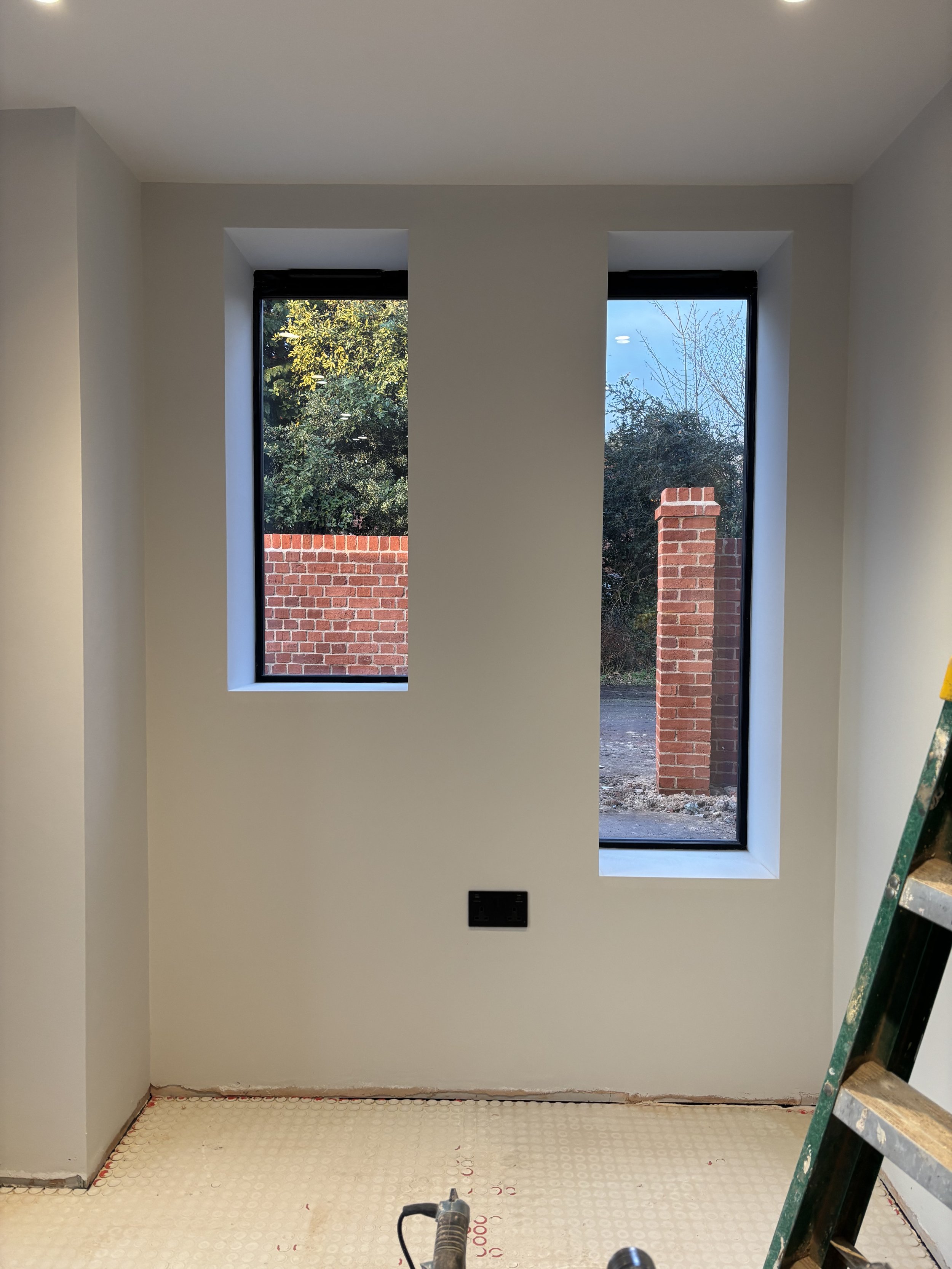 Interior of a room under construction with two tall windows showing a view of a brick wall and trees outside. The floor is unfinished with insulation or underlayment visible. A ladder is on the right side, and tools are on the floor.