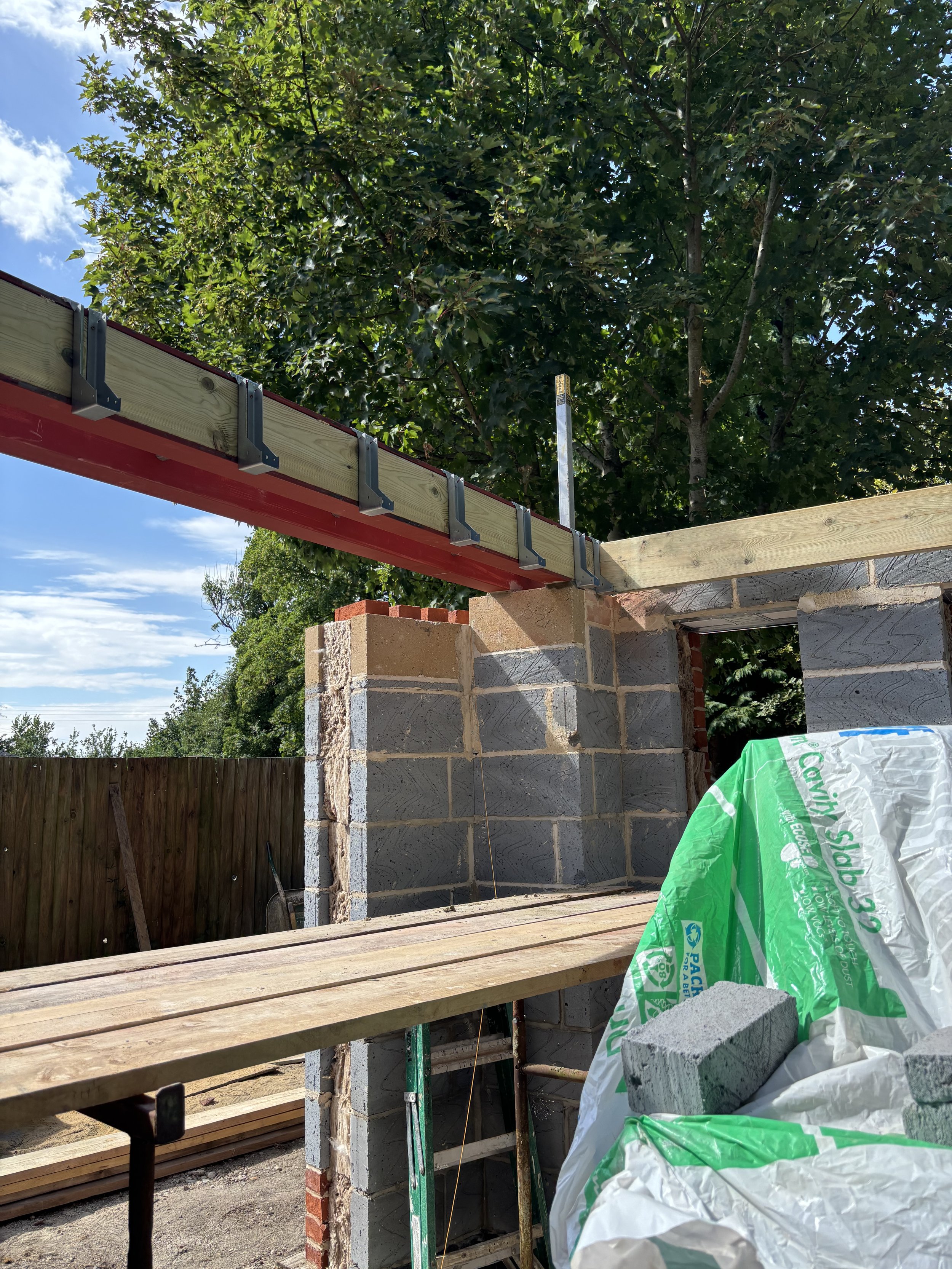 Part of a construction site with a brick wall, wooden beams, a metal ladder, and sacks of construction materials under a clear blue sky with green trees in the background.