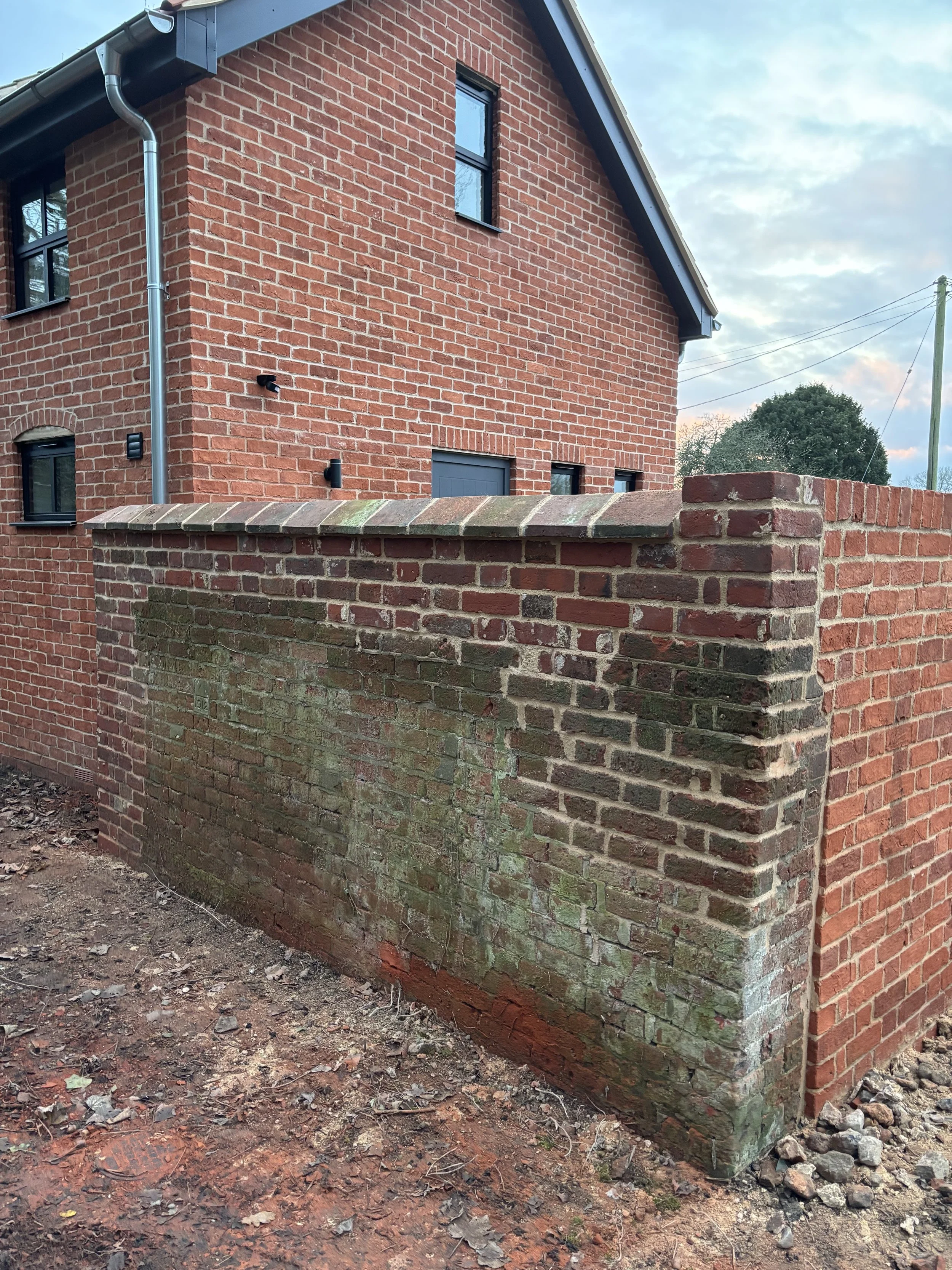 A brick building with a brick wall in the foreground and a larger brick house in the background. The house has a gabled roof, windows, and a dark-colored door. The sky is partly cloudy.