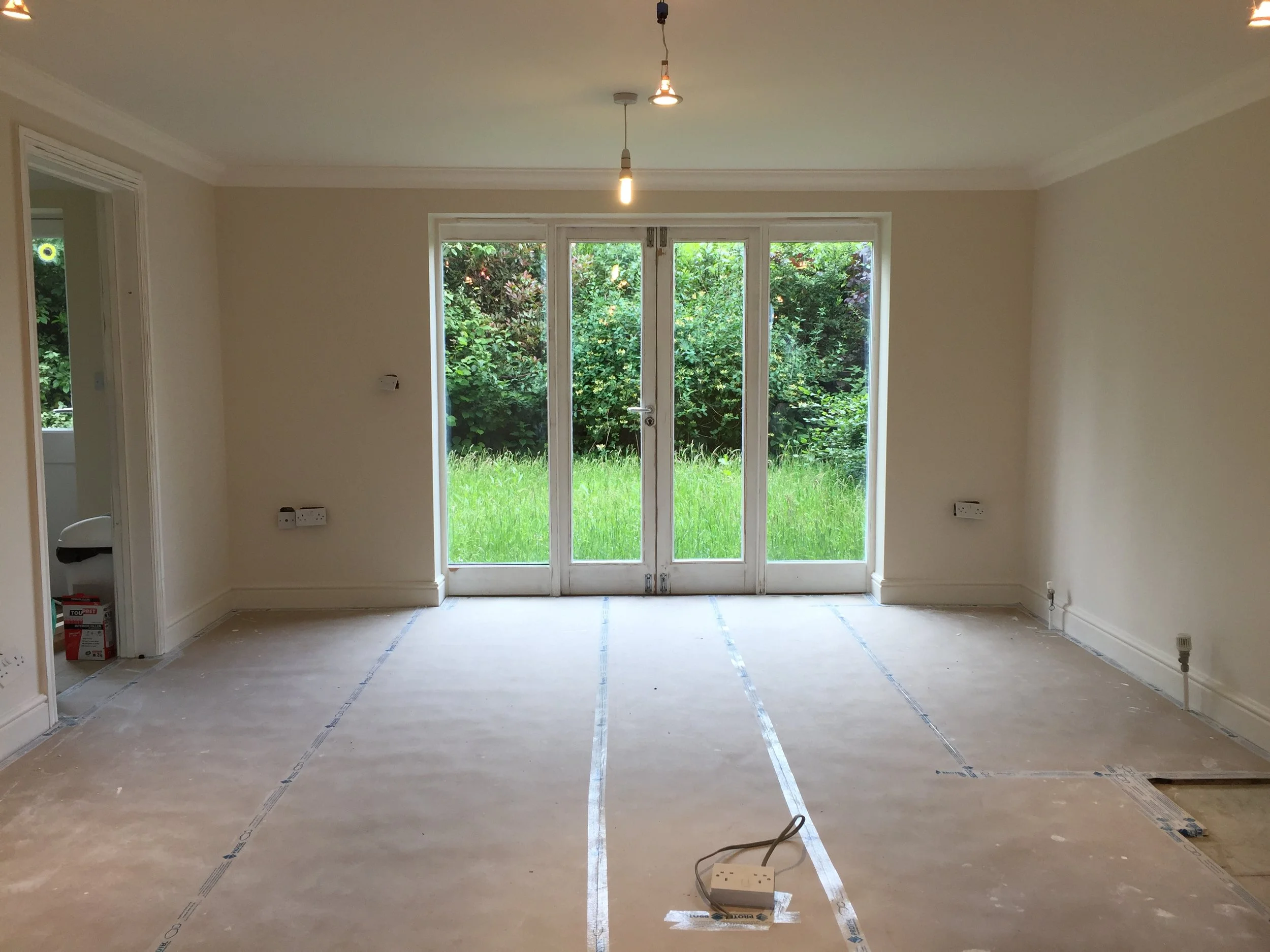 Empty room under renovation with large glass doors to a green garden, light fixtures hanging from the ceiling, electrical outlets with cords on the floor, and construction materials in the corner.