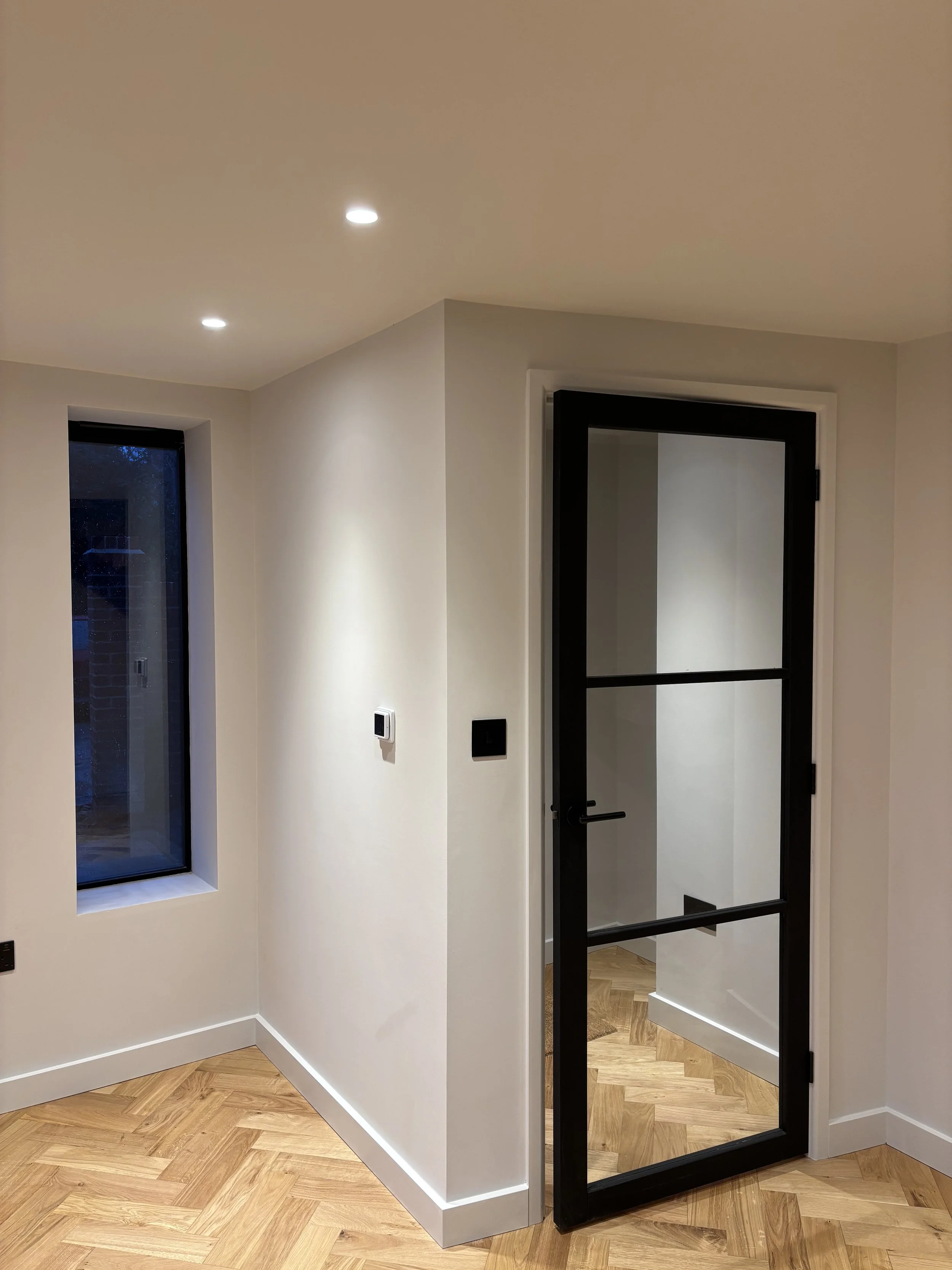 Indoor space with white walls, a small window, and a black-framed glass door. The floor has a herringbone pattern of light wood. There is a thermostat and a black switch on the wall near the door.