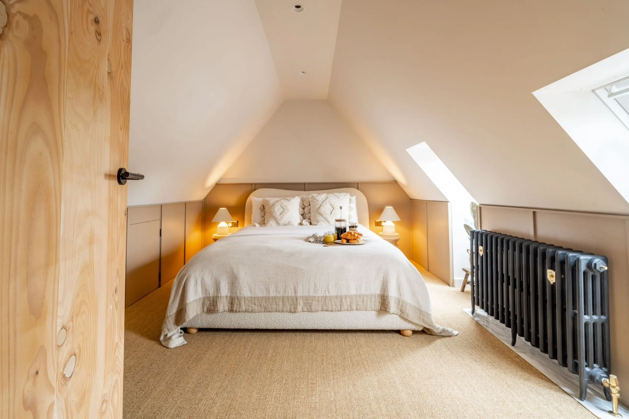 A cozy bedroom with sloped ceilings, a bed with white linens and decorative pillows, bedside lamps, a tray with breakfast on the bed, a wooden door, a window letting in natural light, and a black radiator.